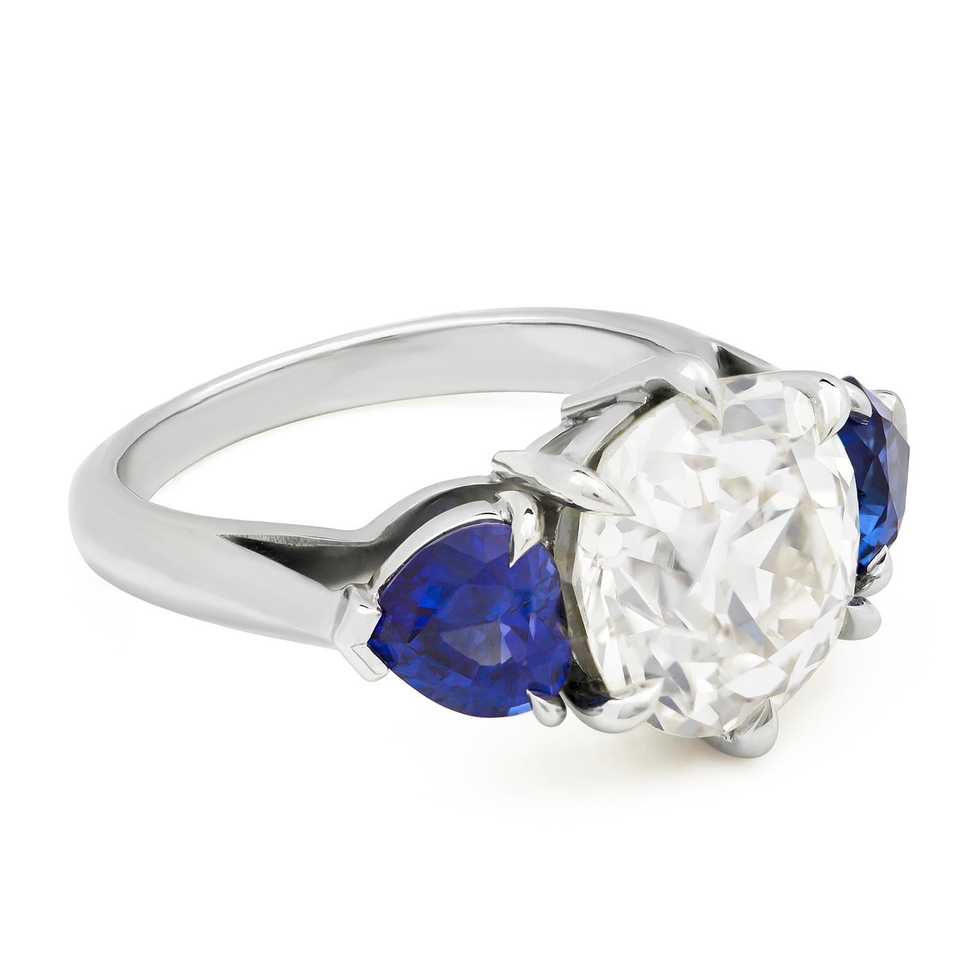 Immortalize the engagement with this gorgeous three-stone engagement ring; features a 4.04 carat old European cut diamond set in a timeless 6 prong setting. Flanking the center stone are two gorgeous and vibrant heart shaped blue sapphires on each
