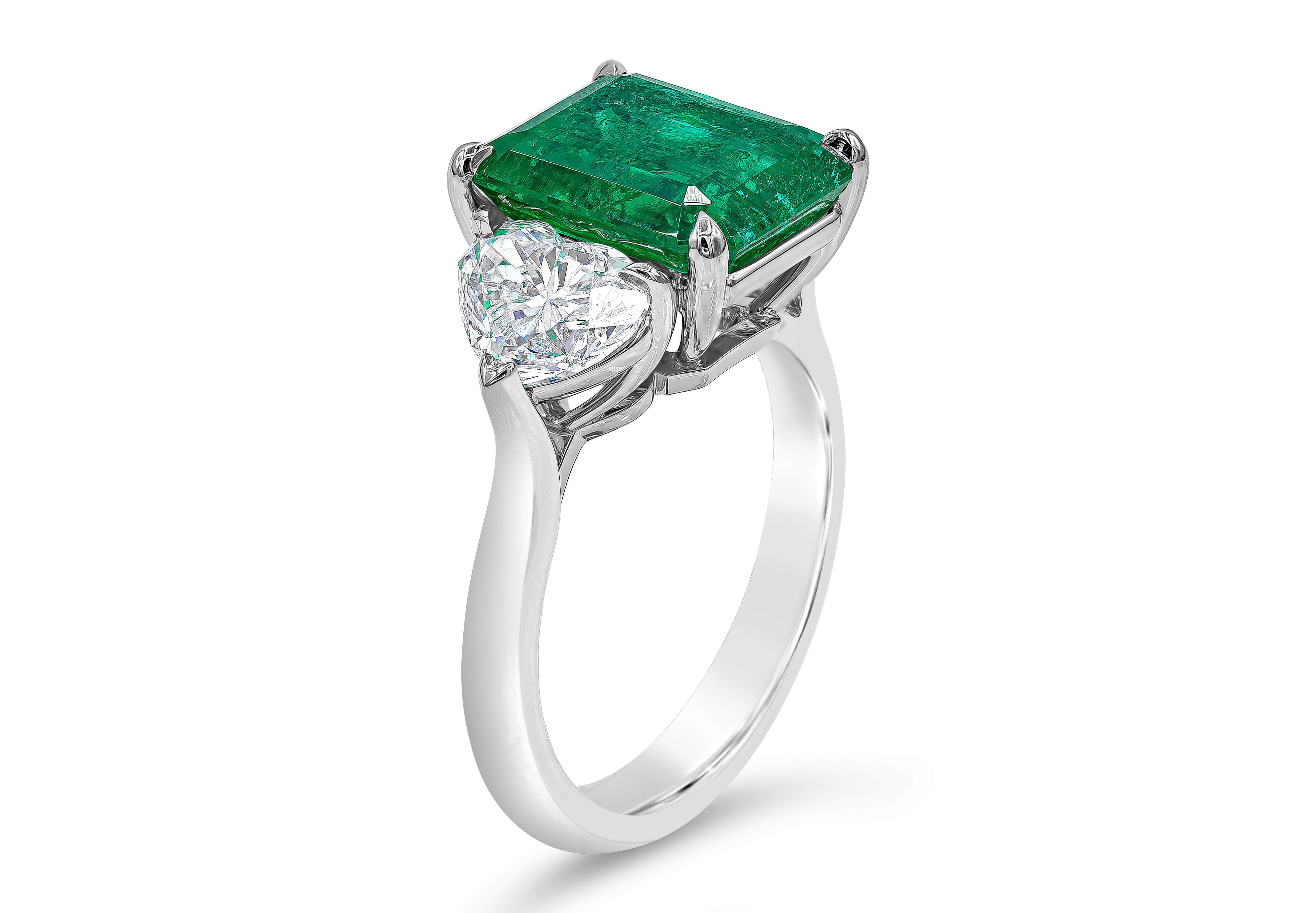 A vibrant and luxurious three-stone engagement ring, showcasing an AGL certified 4.46 carats emerald cut Colombian green emerald, set on a four prong basket setting. Flanked by two brilliant cut heart shape diamonds weighing 2.01 carats total