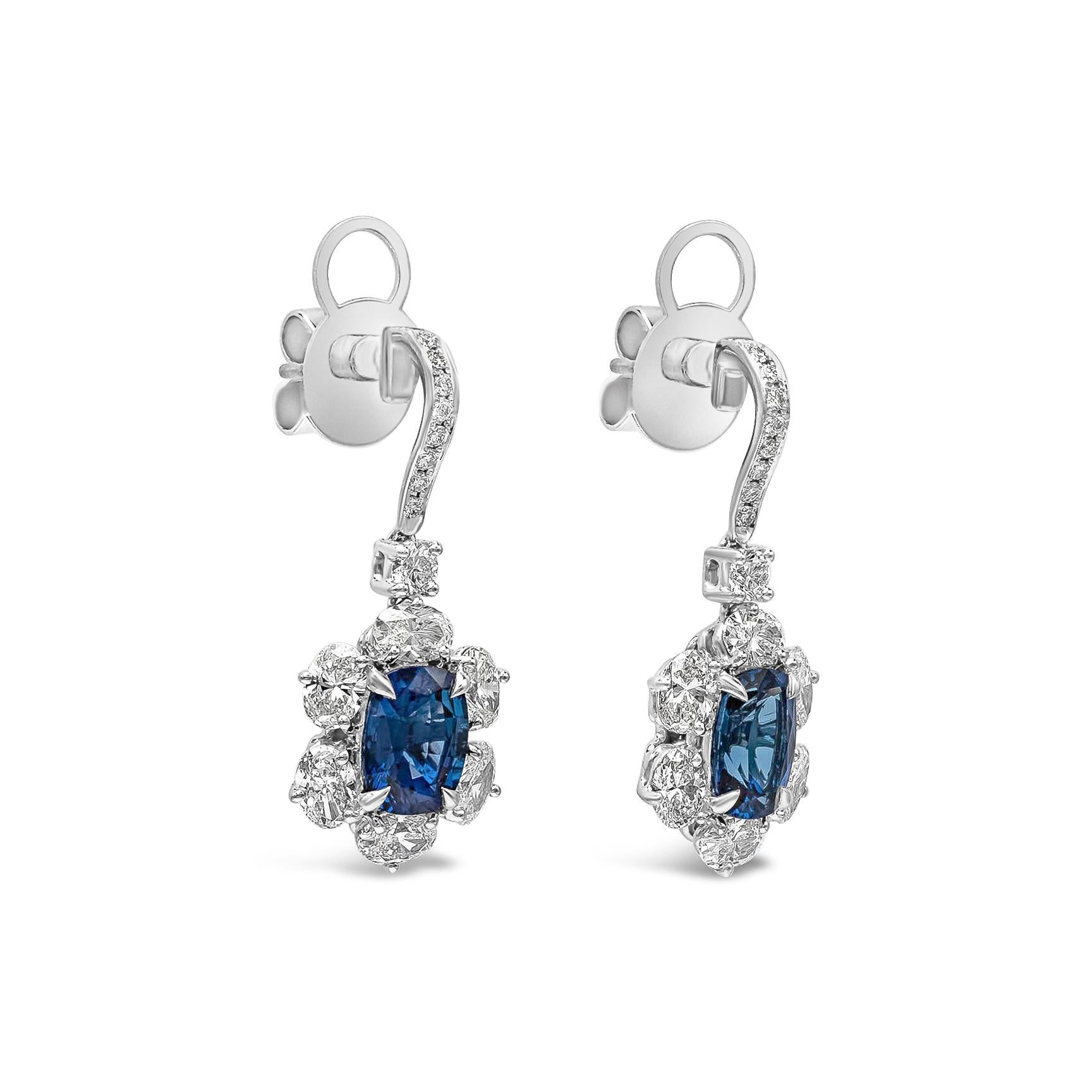 A gorgeous pair of dangling earrings, showcasing vibrant 3.30 total carats of cushion cut blue sapphires, surrounded by oval cut diamonds of 2.50 total carats. Set in a floral-motif design and suspended in a diamond encrusted thin wire made of 18K