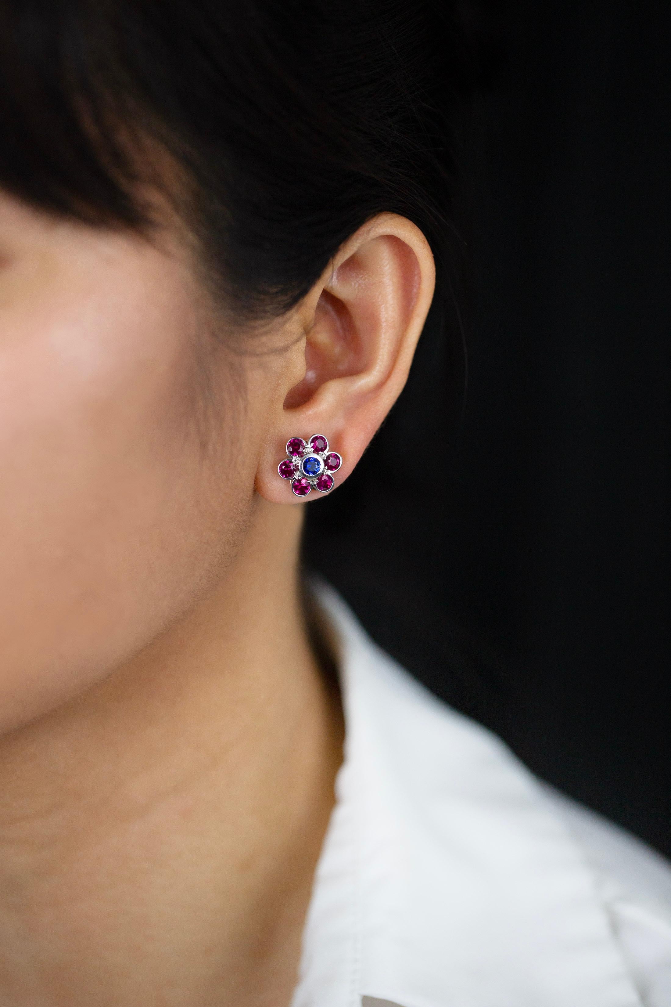 A fashionable pair of stud earrings showcasing a brilliant round cut bezel set blue sapphire in the center weighing 0.55 carats total. Surrounded by six brilliant round rubies weighing 2.52 carats total, arranged in a beautiful floral-motif design.