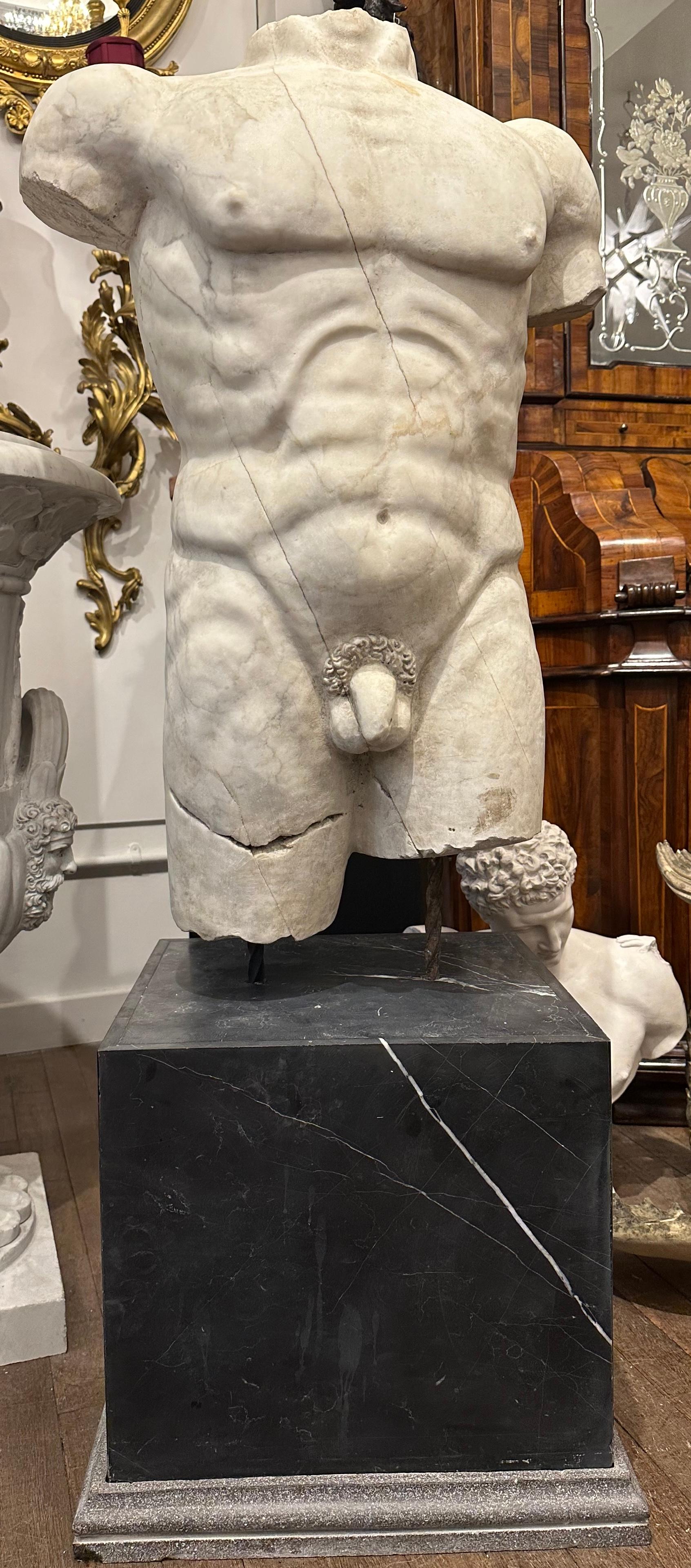A handsomely carved male marble torso on a black marble base. The carving is detailed and realistic, clearly depicting the contours and muscles. The marble has a aged look, matt with veining and some cracks in keeping with its age and the original