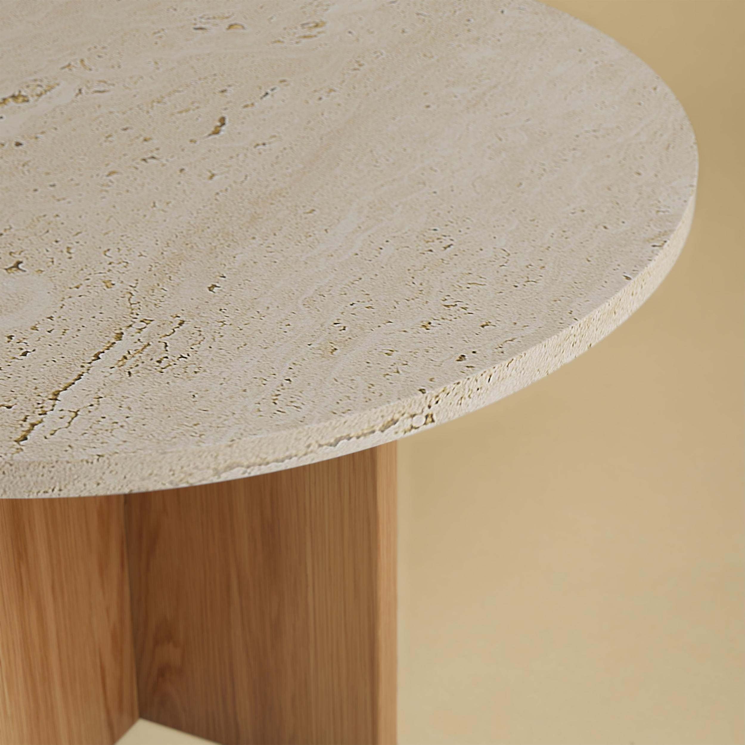 La table basse Tinian est fabriquée avec une base en bois de chêne et un plateau en travertin romain. Le plateau est circulaire et mesure 60 cm de diamètre, tandis que la base est obtenue en collant des planches de chêne perpendiculairement les unes