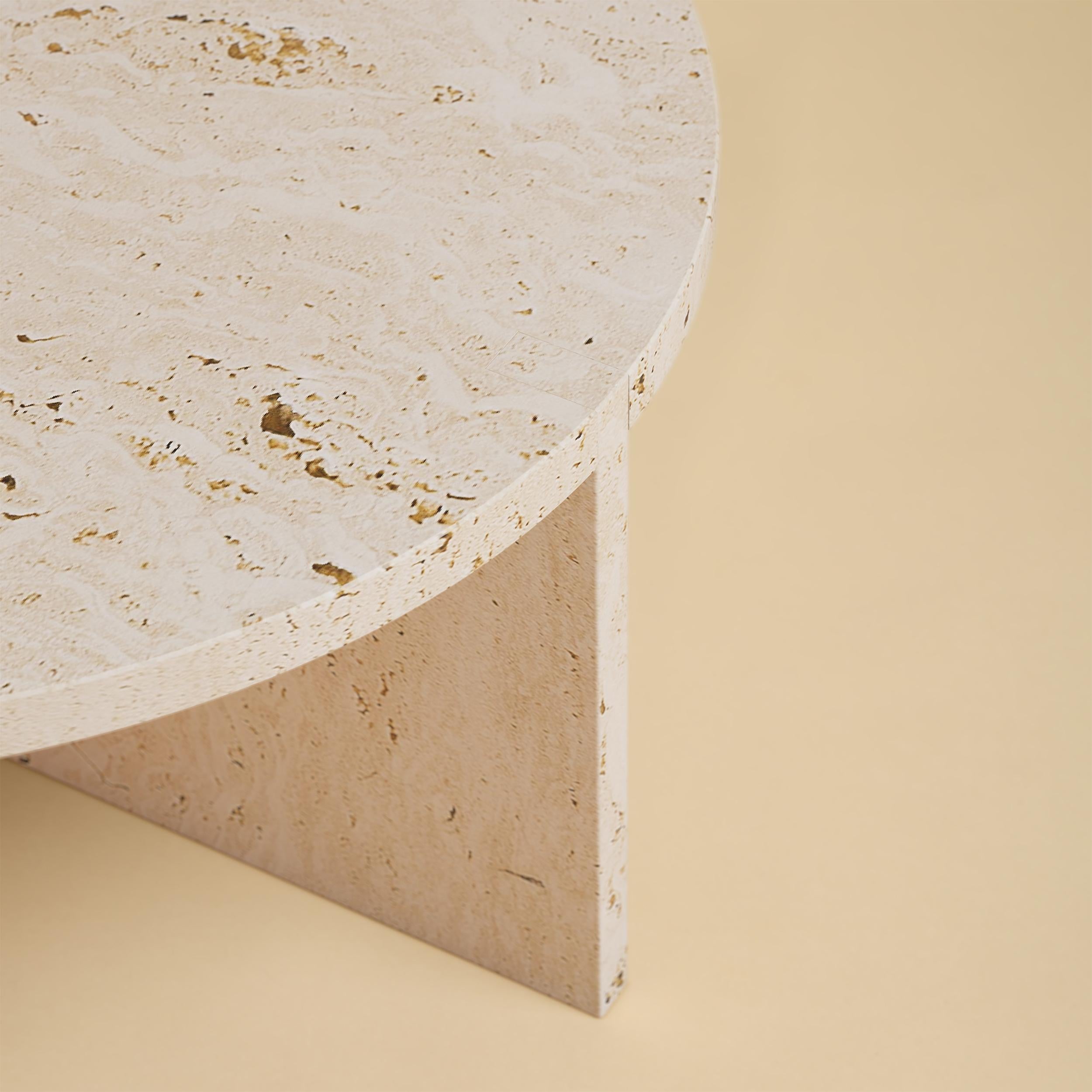 La table basse Hashi est entièrement réalisée en travertin romain d'Italie. Le plateau est circulaire et mesure 60 cm de diamètre. Les pieds sont constitués de deux plaques de marbre dont une partie est incrustée sur le plateau, ce qui constitue un
