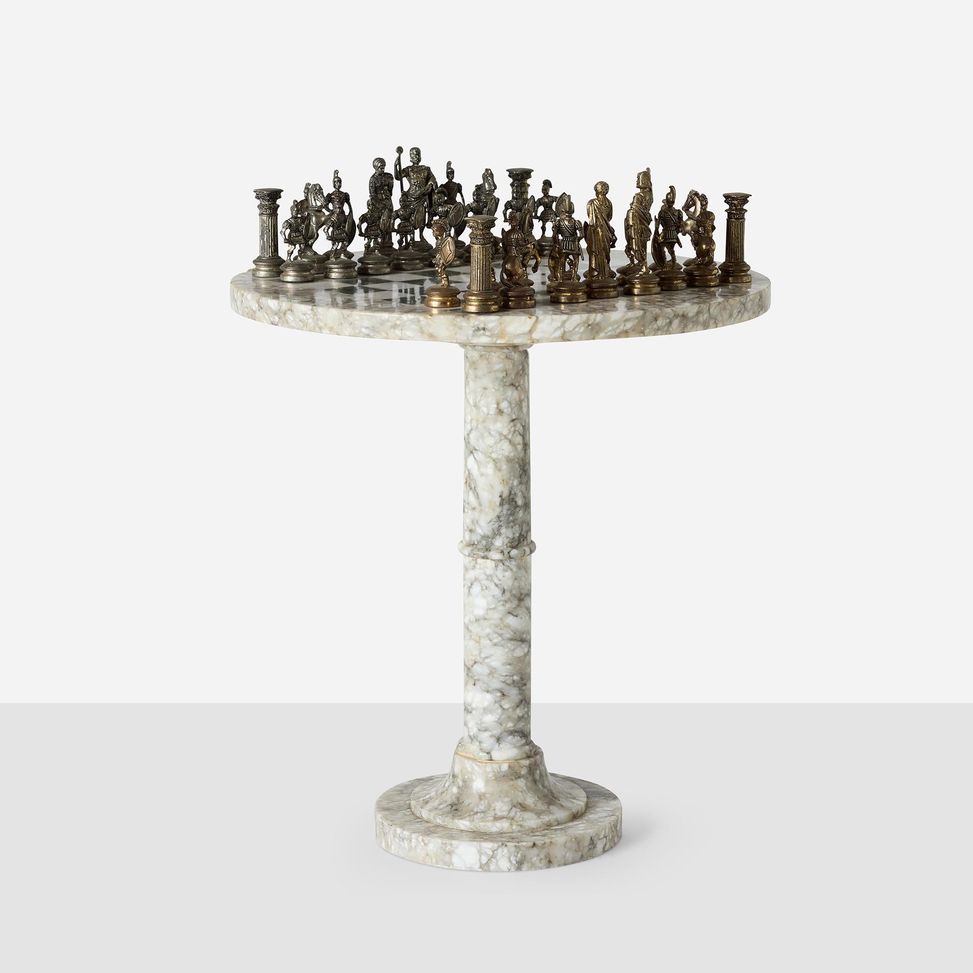 Cast metal chess set depicting Romans versus Greeks on custom marble pedestal table. 

The chess pieces feature detailed classical warrior figures, with one side representing Roman soldiers and the other Greek soldiers. Includes armored warriors,