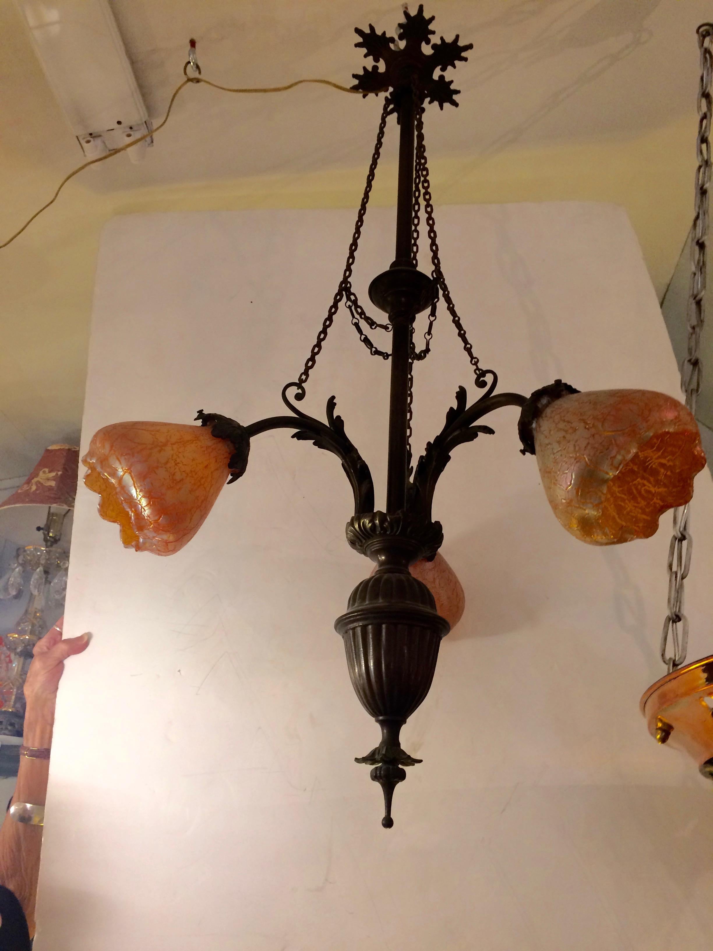 Gorgeous bronze and brass Art Nouveau light fixture having an elongated structure, lovely wheel-like ceiling cap and three arms displaying soft tangerine colored crinkle glass and scalloped shades. Original finish, rewired, 40 watt each max.