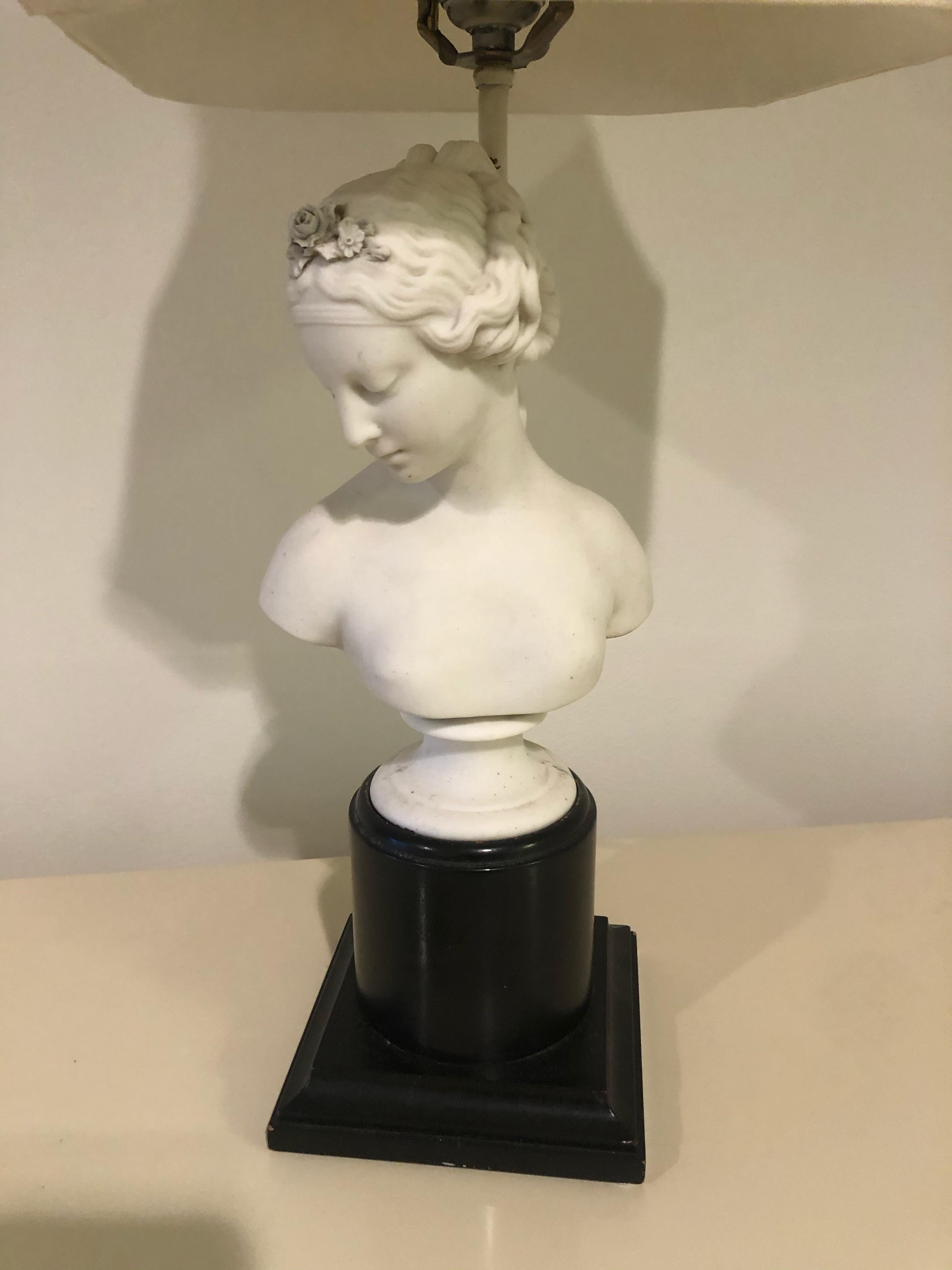 A lovely vintage female plaster bust sculpture converted into a striking table lamp having off white silk shade topped with a wooden finial. White paster bust rests on a black wood plinth.
 