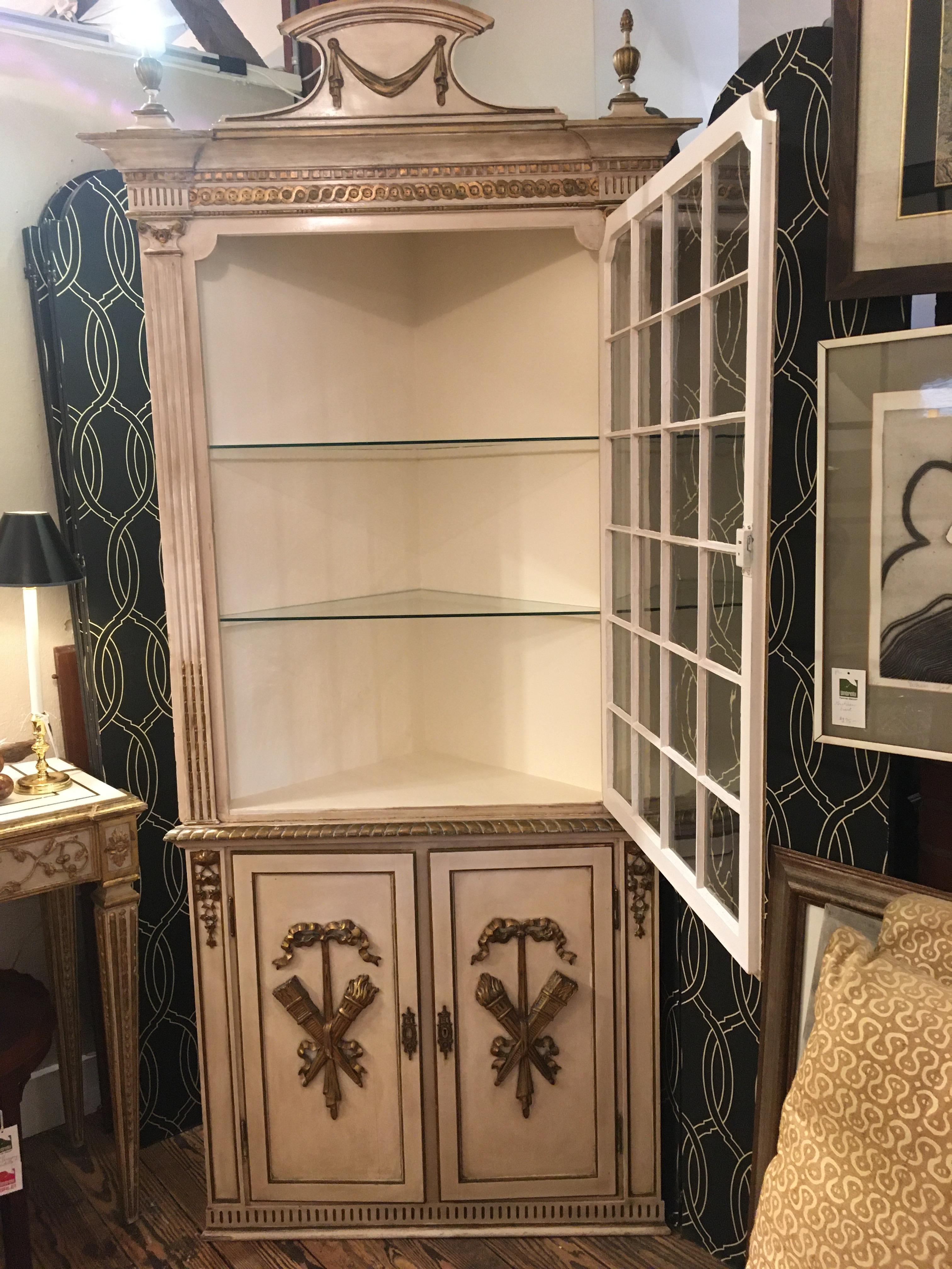 A glamorous French antique carved wood corner cupboard painted a creamy white having giltwood embellishments including swag and finials at the top and torches with bows on the panel doors below. Original glass is in the top single door that opens to