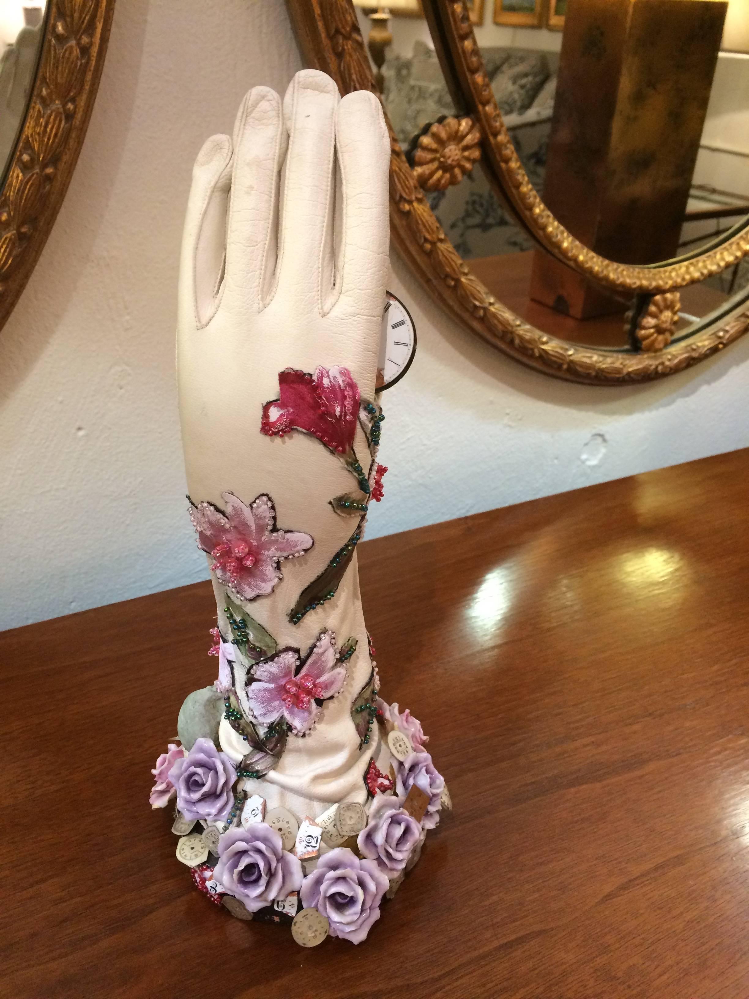 Nachdenkliche Mischtechnik-Skulptur einer Hand mit Lederhandschuhen, die mit Porzellanblumen, floralen Perlenverzierungen und einem Totenkopf verziert ist und provokativ ein rosafarbenes Ziffernblatt hält, das den Lauf der Zeit andeutet.