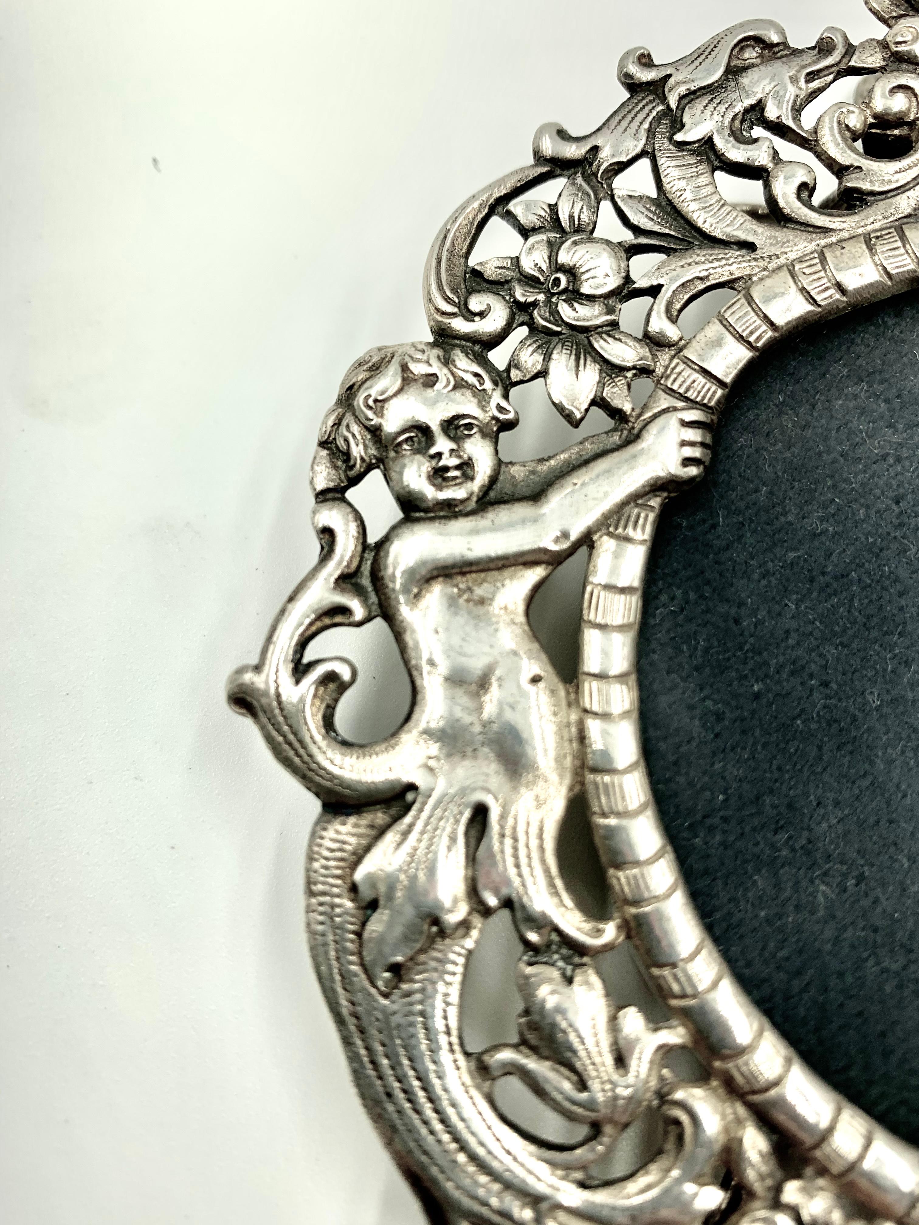 Fabulous European antique silver picture frame with a pair of angels on each side holding the oval of the frame amidst floral scroll decorations with a crest featuring a pair of dolphins flanking a central footed centerpiece filled with exotic