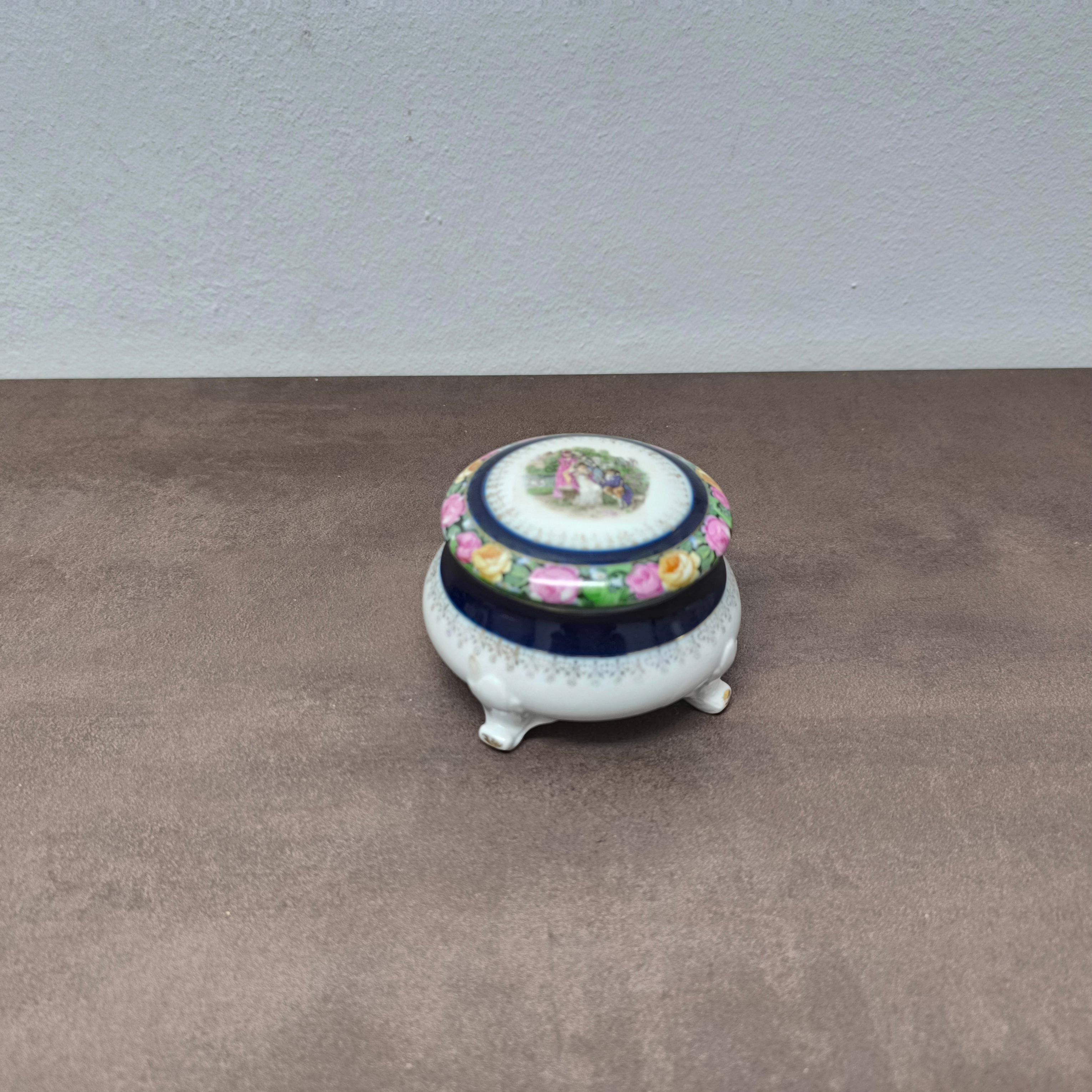 Boîte à bibelots romantique en porcelaine avec scène de jardin, France, milieu du 20e siècle
Charmante boîte à bibelots en porcelaine peinte à la main, probablement française, datant du milieu du XXe siècle. Mesurant 10 cm de diamètre et 7 cm de