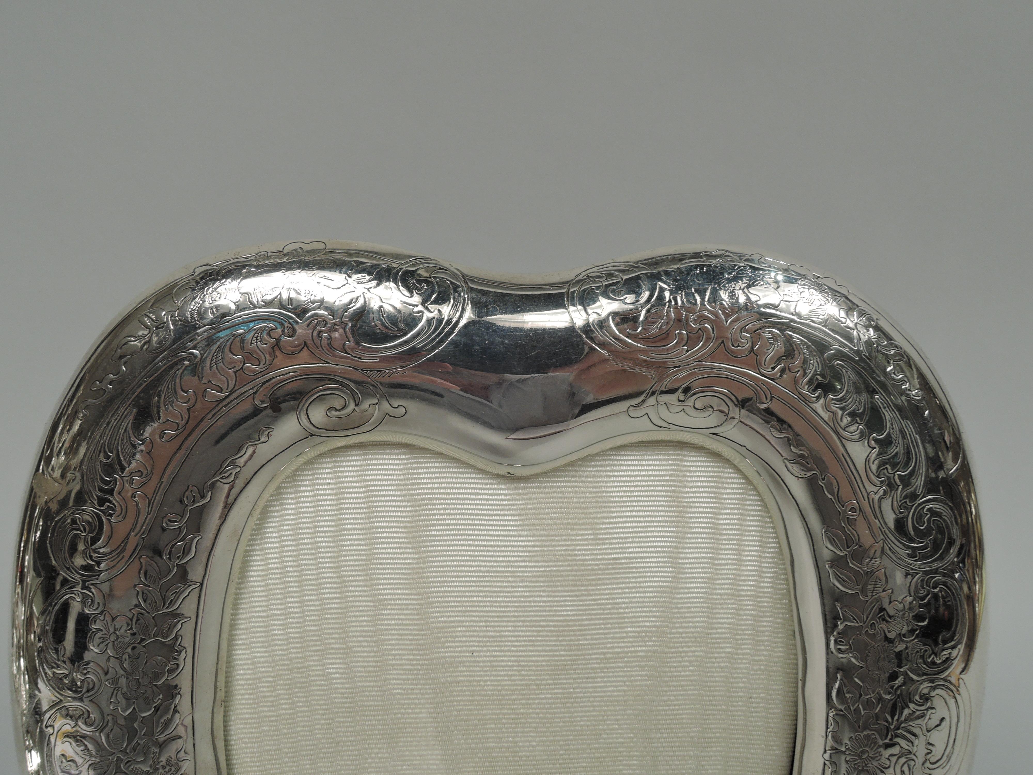 Romantic Edwardian Art Nouveau sterling silver frame. Made by Tiffany & Co. in New York. Heart-shaped window in same and curved surround flowing into bracket feet. Acid-etched leafing scrollwork and flowers. At top shaped cartouche vacant for