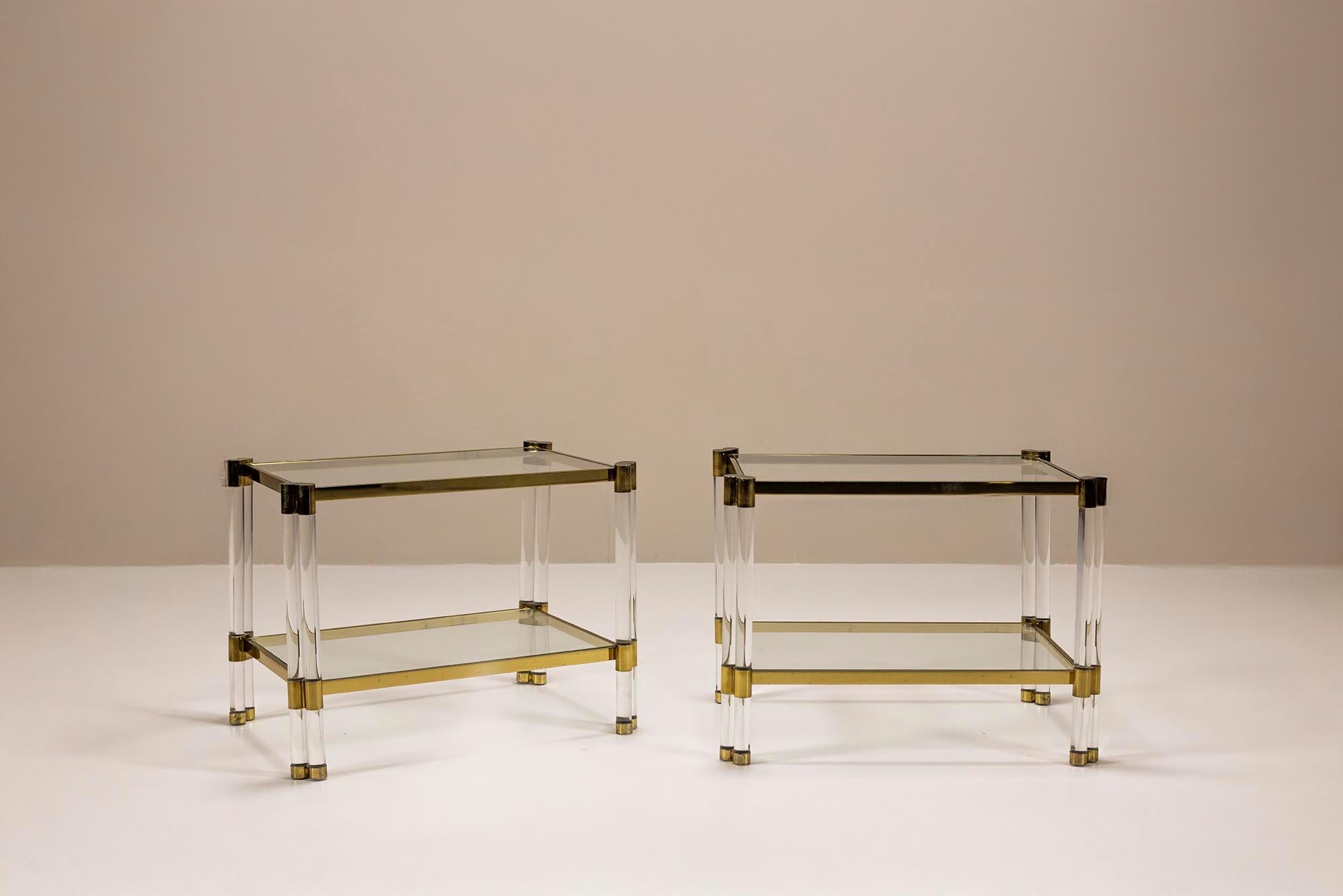 This pair of side tables is composed of thick Lucite legs, transparent glass shelving, and a linear brass frame. The twin cylindrical legs are joined by heart-shaped brass collars at each corner, a distinctive design element often attributed to