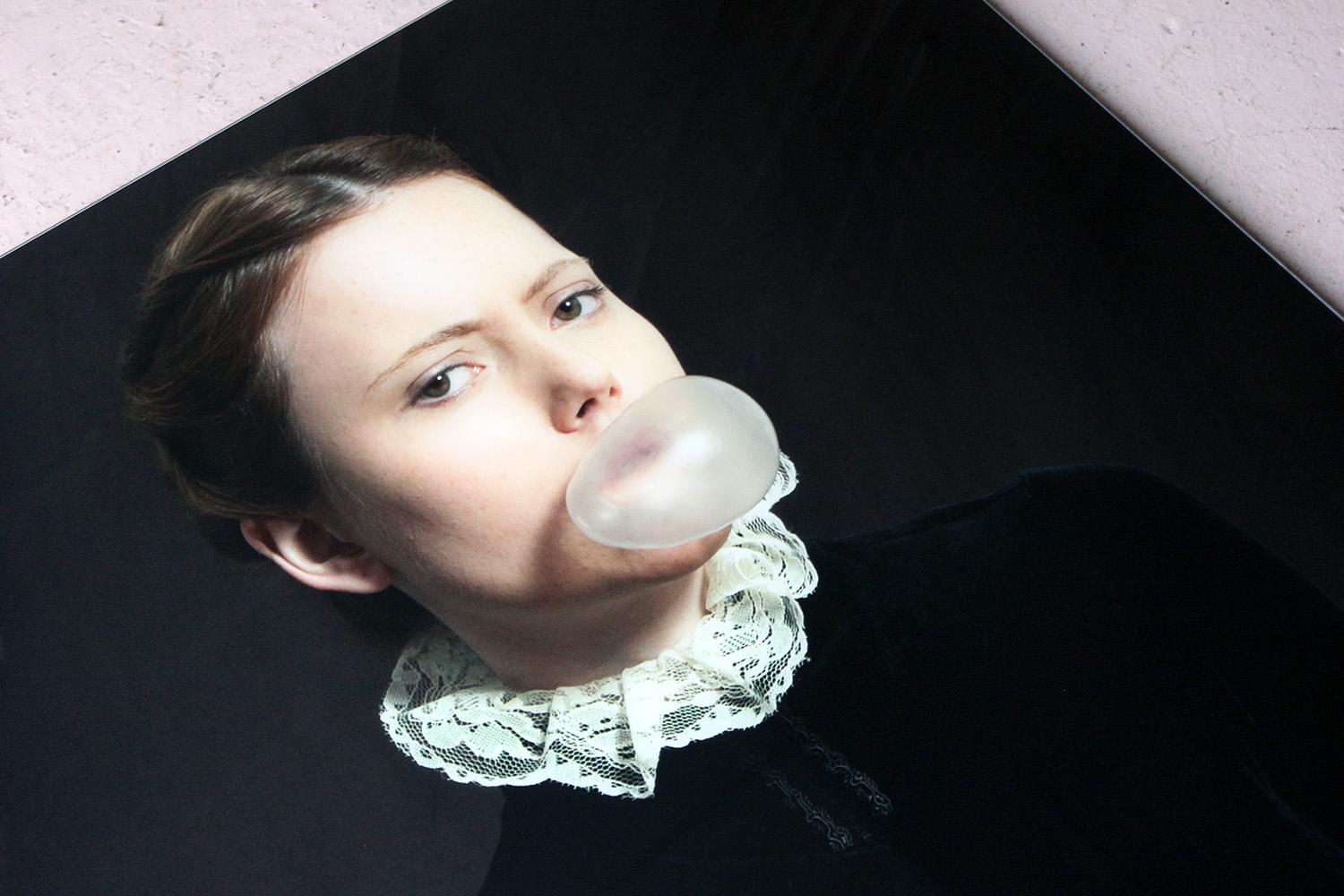 Die in einem Rahmen aus Aluminium und Acrylglas eingefasste Großdruckfotografie von Romina Ressia aus dem Jahr 2014 mit dem Titel Bubblegum zeigt eine junge Frau in halber Körpergröße aus der Renaissance, die sitzend Kaugummi bläst und dabei eine