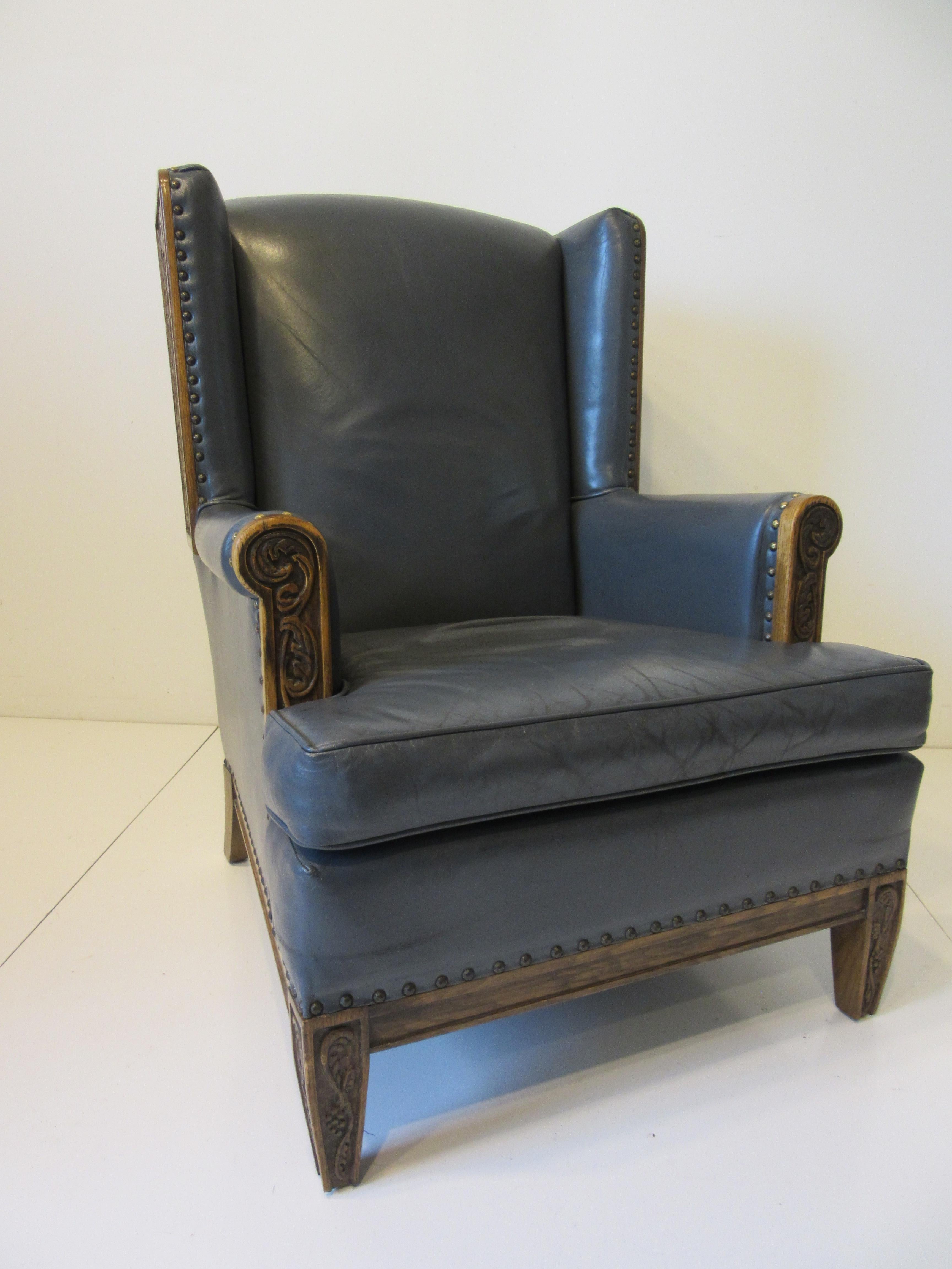 A medium sized gray blue leather wingback lounge chair with oak wood carved wings depicting grapes and vines with the front legs and arm area having the same motif. Heavy leather with antiqued brass stud detail defines the border of the leather in a