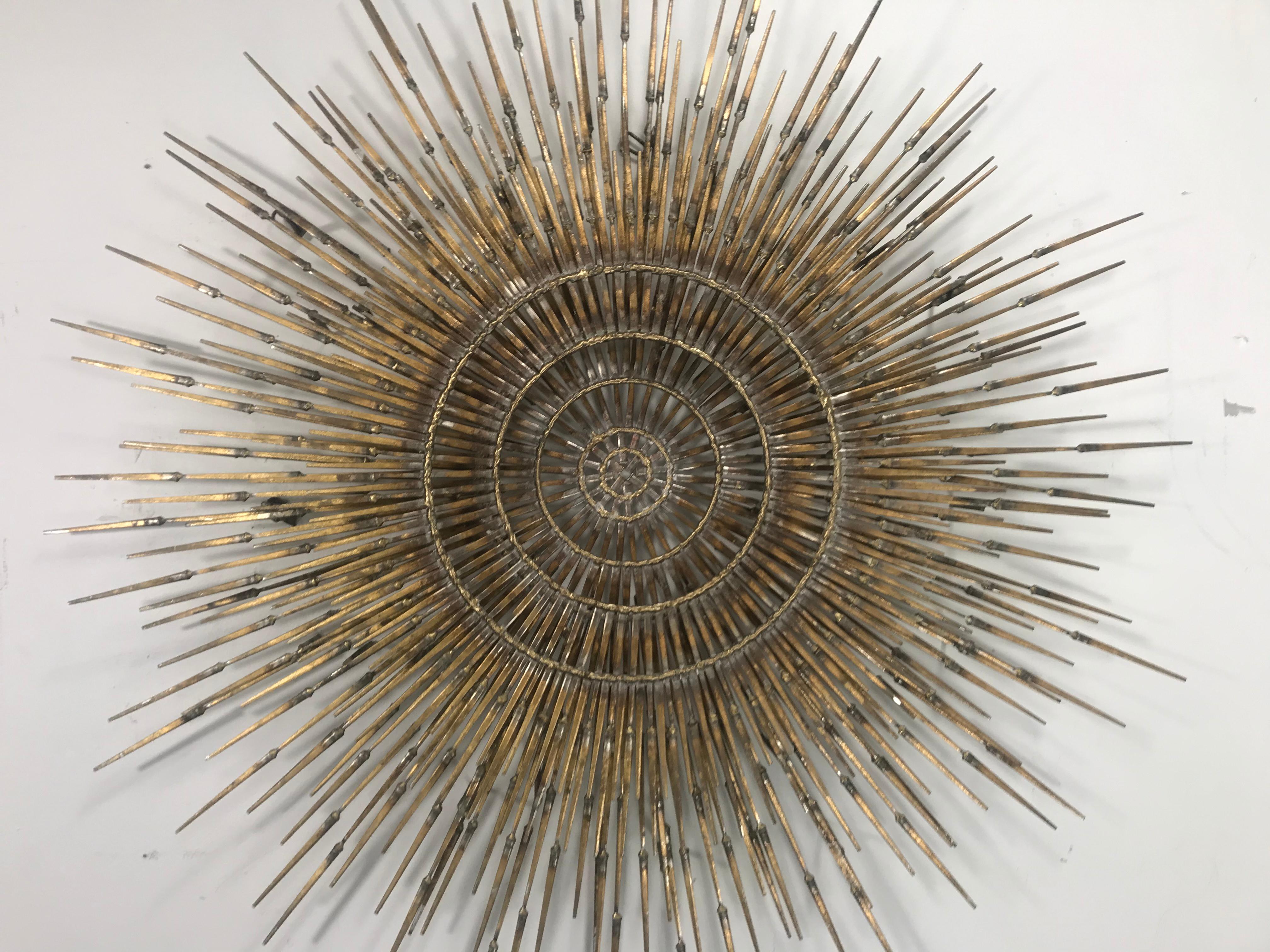 A large 4 layer Brutalist sunburst with starburst overlay nail art wall sculpture by Ron Schmidt.

Very uncommon multiplanar design with brass welded iron nail rays radiating sunburst-style from a central brass ring that floats in front of an open