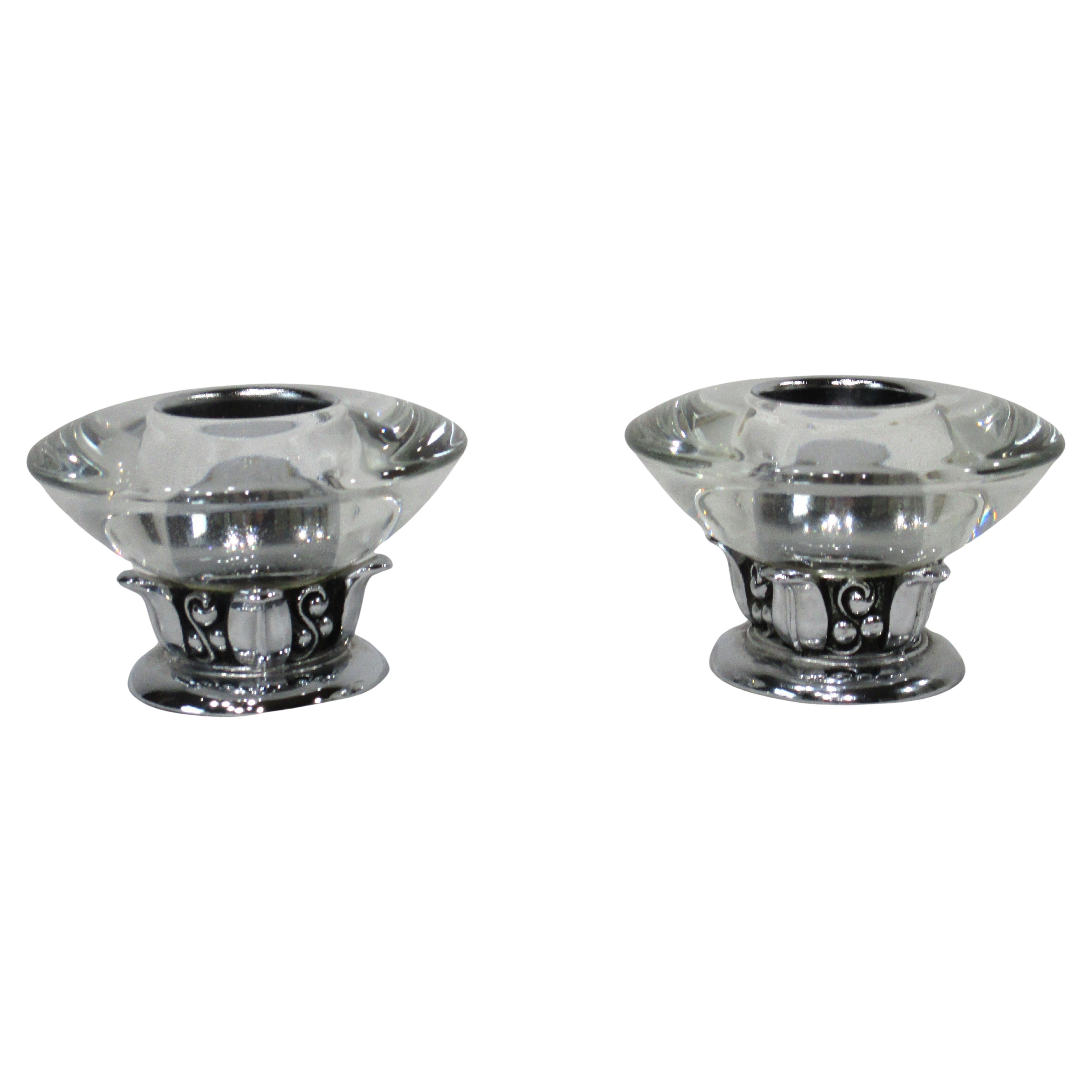 A pair of candlesticks with molded glass tops and heavy chrome bases with leaf and vine designs around the collar . Manufactured by the Ronson company noted for their lighters and smoking items .