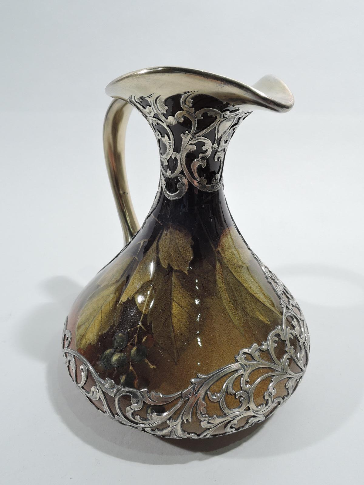 Art Nouveau Craftsman glazed silver overlay pitcher. Made by Rookwood Pottery in Cincinnati in 1894. Conical body with crimped trefoil mouth and scroll handle. Fruiting grapevine with overlapping leaves in the greenery-yallery palette and blue