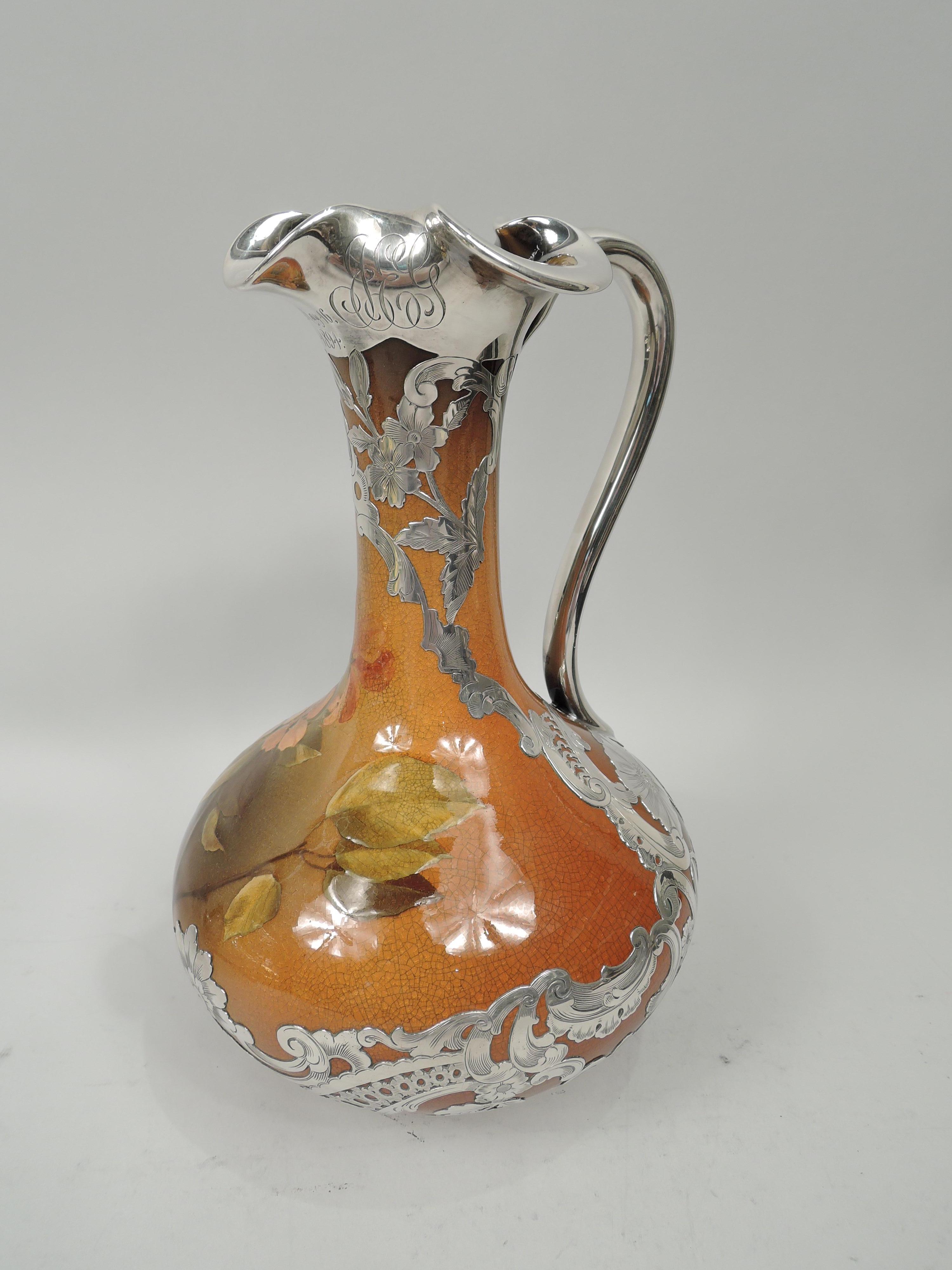 Art Nouveau Craftsman glazed earthenware wine ewer with engraved silver overlay. Made by Rookwood Pottery in Cincinnati in 1893. Bellied bowl, cylindrical neck, silver looping scroll handle, and lobed trefoil mouth in silver collar engraved with the