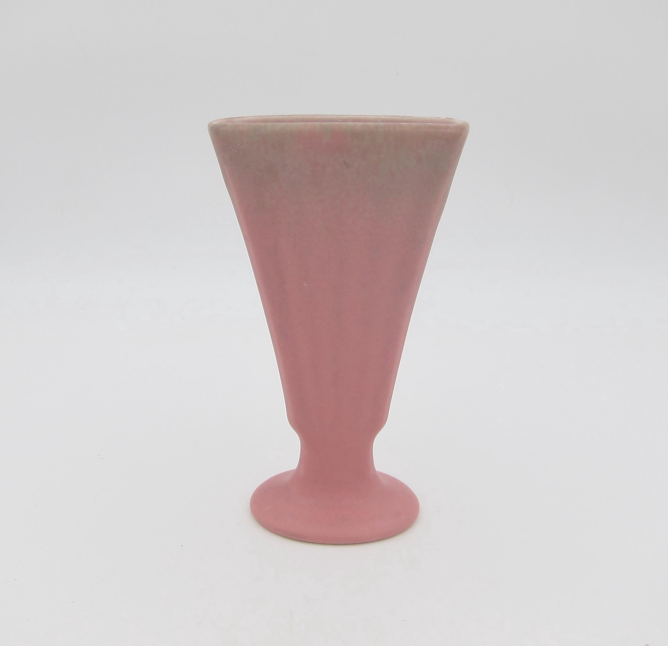 An American Rookwood Pottery production vase date marked for 1927. The American art pottery vase flares outward in a fan shape with subtle ribbed detailing resting atop an oval foot. The vessel's matte glaze is a velvety dusty rose-pink with mottled