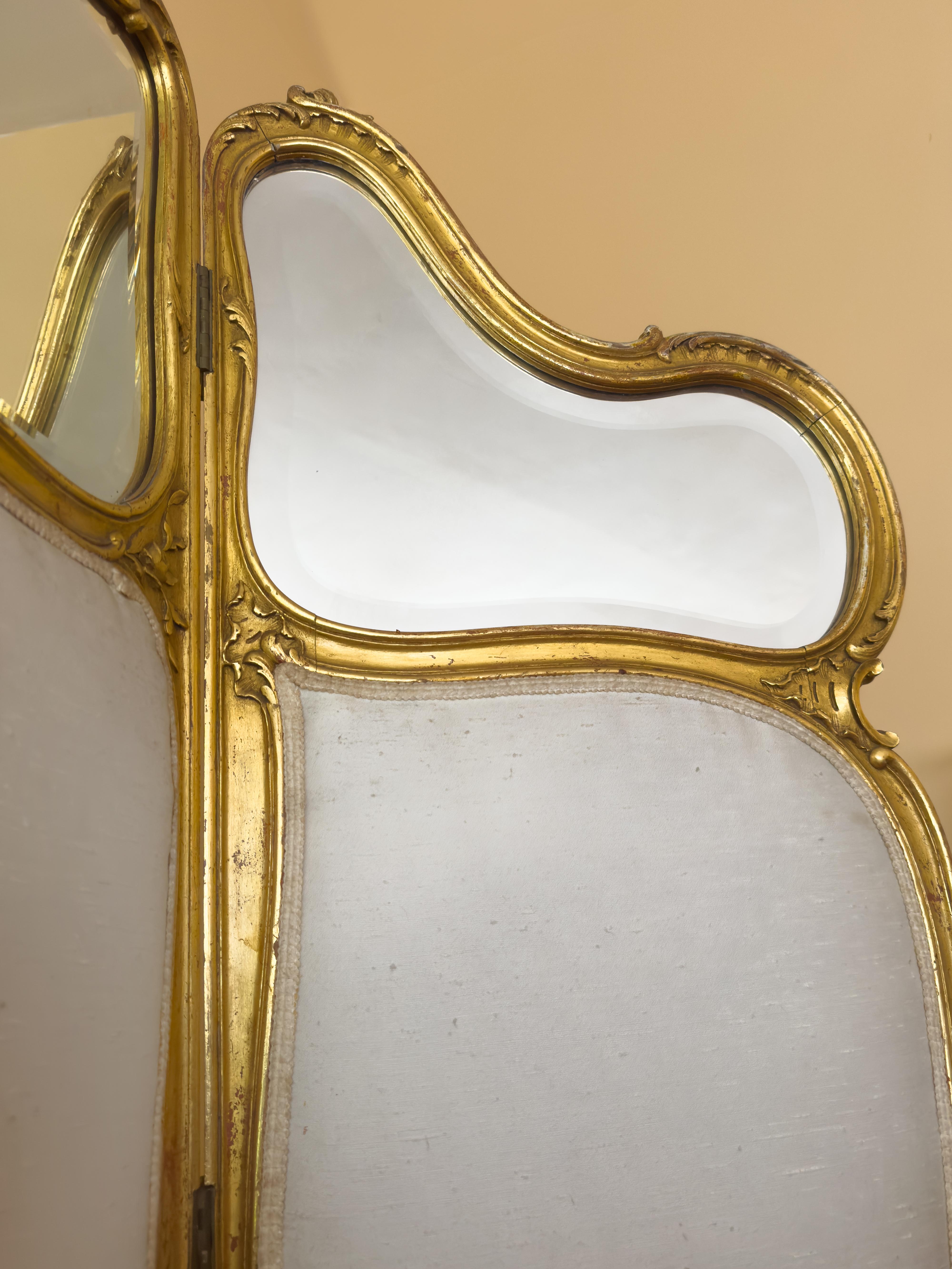 This elegant Louis XV style three-panel room divider from the 20th century is crafted in gilded wood, showcasing graceful curves and classic decorative detailing. Each panel features a mirror surface at the top and is fabric-covered at the bottom