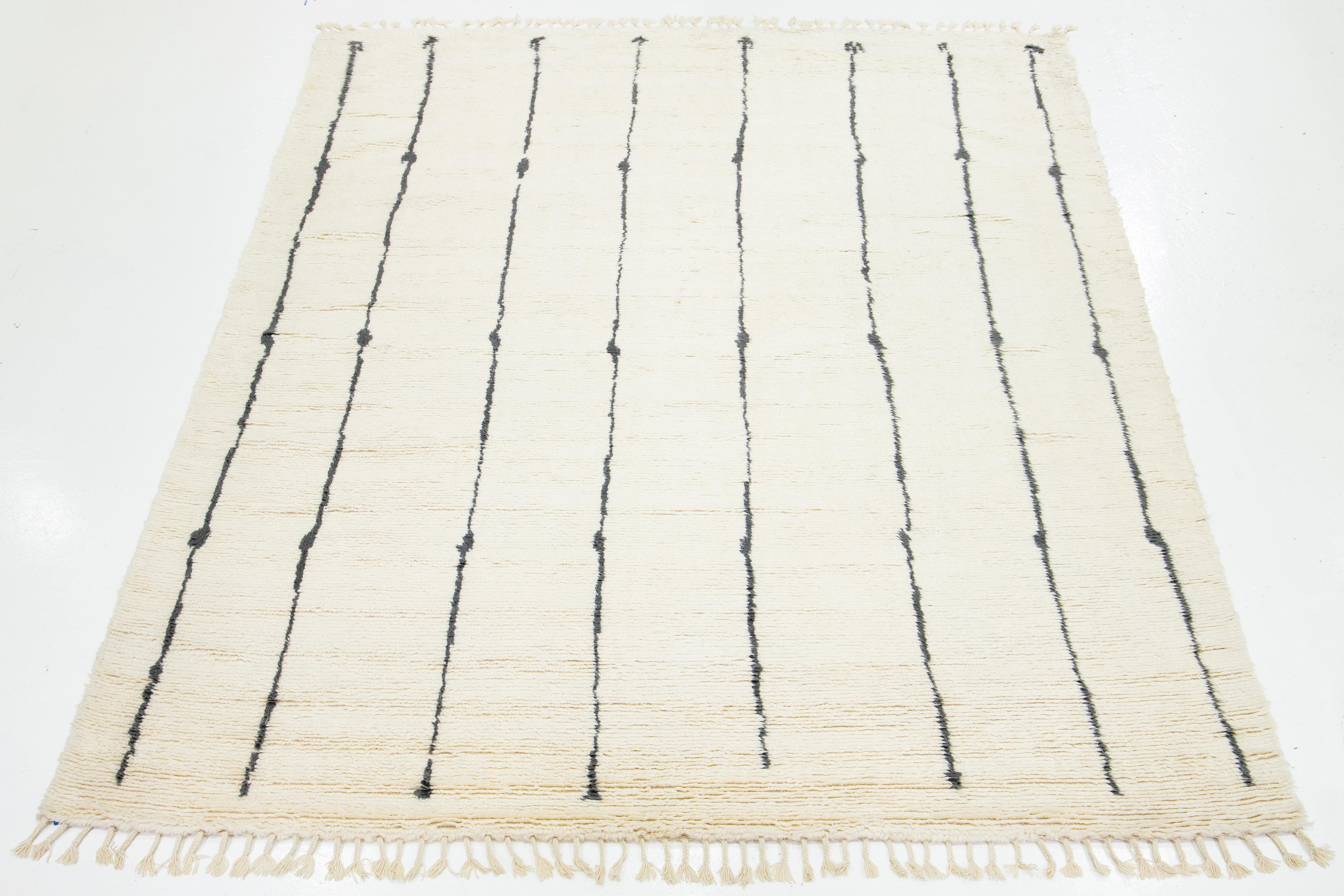 Dieser moderne, handgeknüpfte Wollteppich im marokkanischen Stil zeichnet sich durch ein elfenbeinfarbenes Feld, Fransen und ein plüschiges, atemberaubendes schwarzes Liniendesign aus.

Dieser Teppich misst 10'1