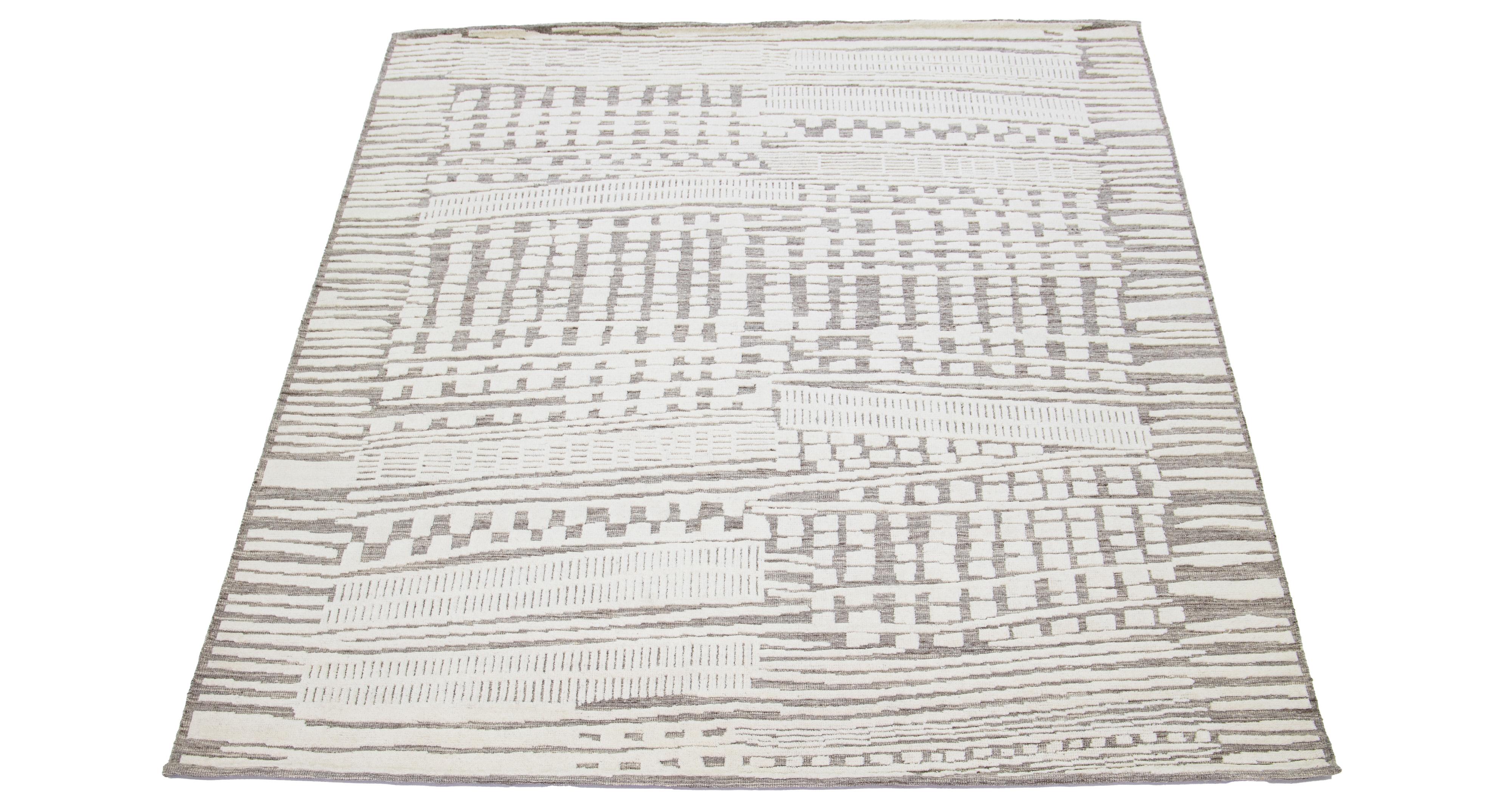 Dieser handgeknüpfte Wollteppich zeigt ein modernes, marokkanisch inspiriertes Motiv, das dezente Beigetöne vor einem kräftigen grauen Hintergrund hervorhebt und so ein fesselndes abstraktes Design schafft.

Dieser Teppich misst 8'1