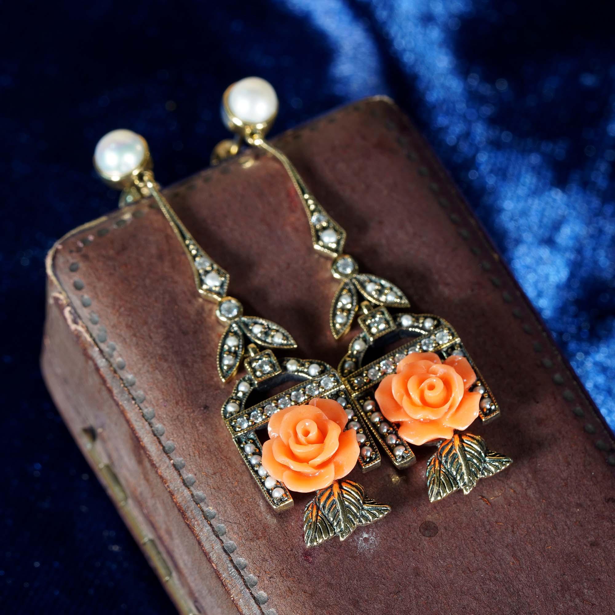 A delicate and feminine pair of Victorian era style carved coral rose drop earrings made in oxidized 9k yellow gold. The natural coral displays a warm salmon color, hanging in a pearl and diamond frame with the leaves beneath. A beautiful piece to