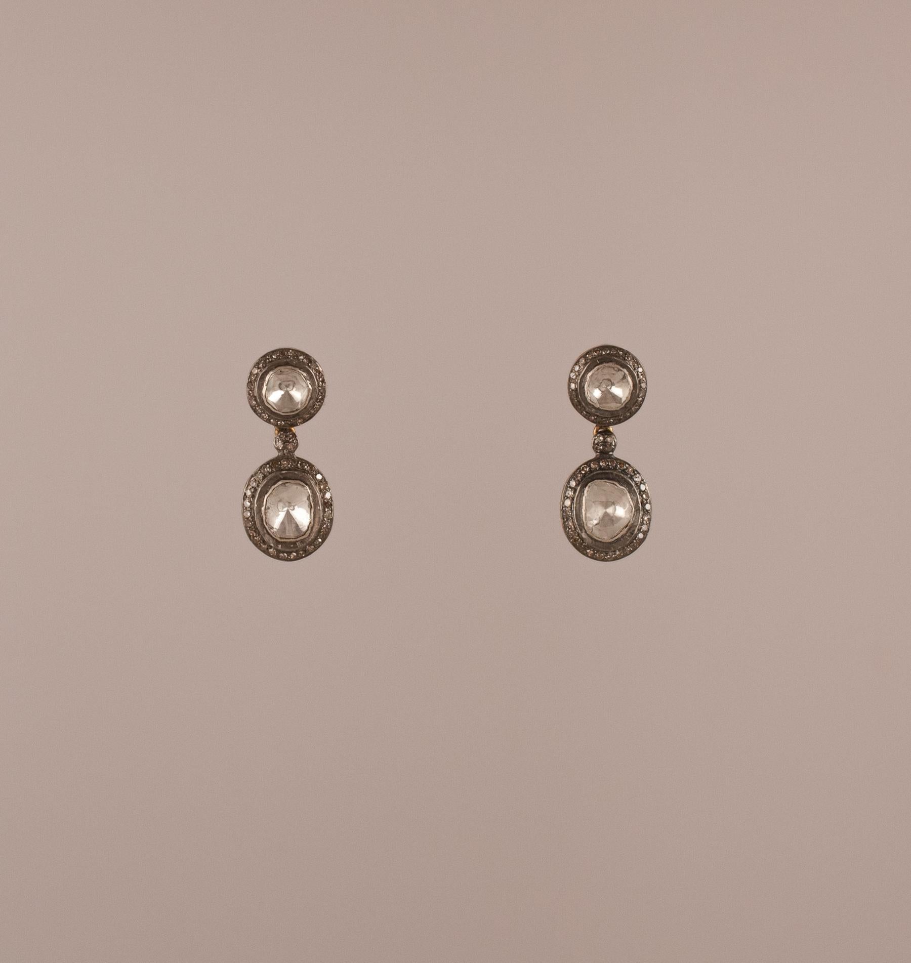 These two-tiered rose cut diamond earrings feature a circular stone on top and an asymmetrical dangling stone beneath, both in a traditional Indian foil-backed setting and encircled by diamonds set in sterling silver. The back of the earrings are an