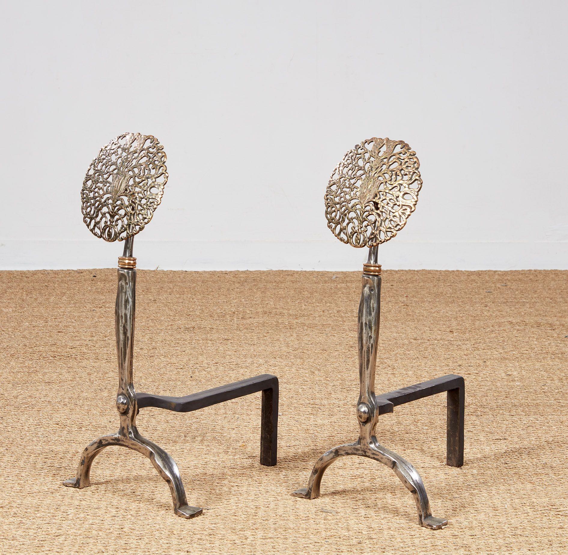 A pair of impressive andirons with large elaborately cast and hand hammered pierced brass rose heads mounted on polished shaped steel shafts with brass collars and with integral arched hoop legs.  Fine quality craftsmanship - possibly Rife Loth,