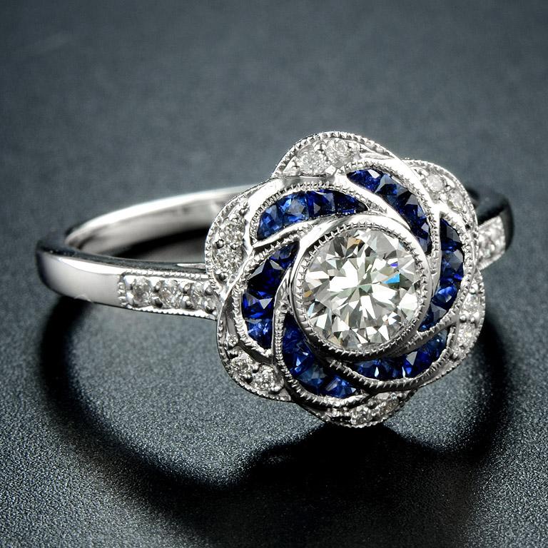 Artistry and sophistication come together in a flower ring with sapphires and diamonds from the Art Deco era. The ring features a round shape center which you can place any round diamond on it in a bezel setting surrounded by calibre French Cut