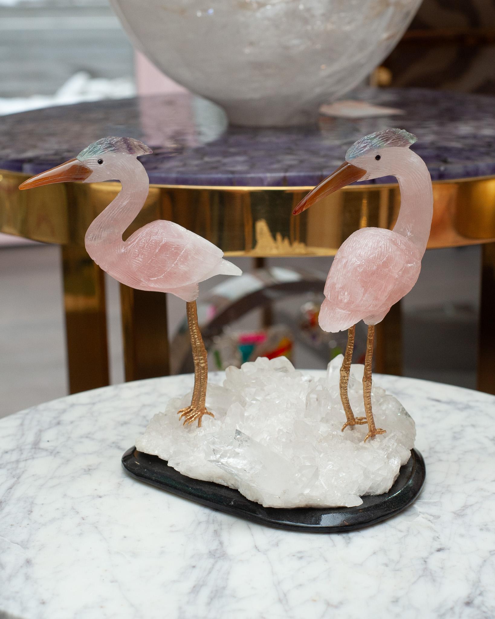 A beautiful hand carved semi precious rose quartz stone crane pair, with carnelian beaks, fluorite crests, and brass legs. The pair of cranes are mounted on a rock crystal quartz mineral specimen on a black marble base. These exotic birds are a