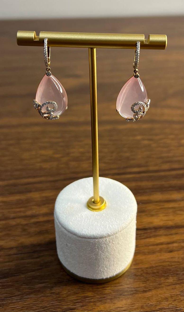 These elegant earrings feature teardrop-shaped pink rose quartz, delicately adorned with intricate, swirling diamond accents of G/VS Quality. The gemstones are suspended from a sleek, diamond-encrusted hoop, adding a touch of sophistication and