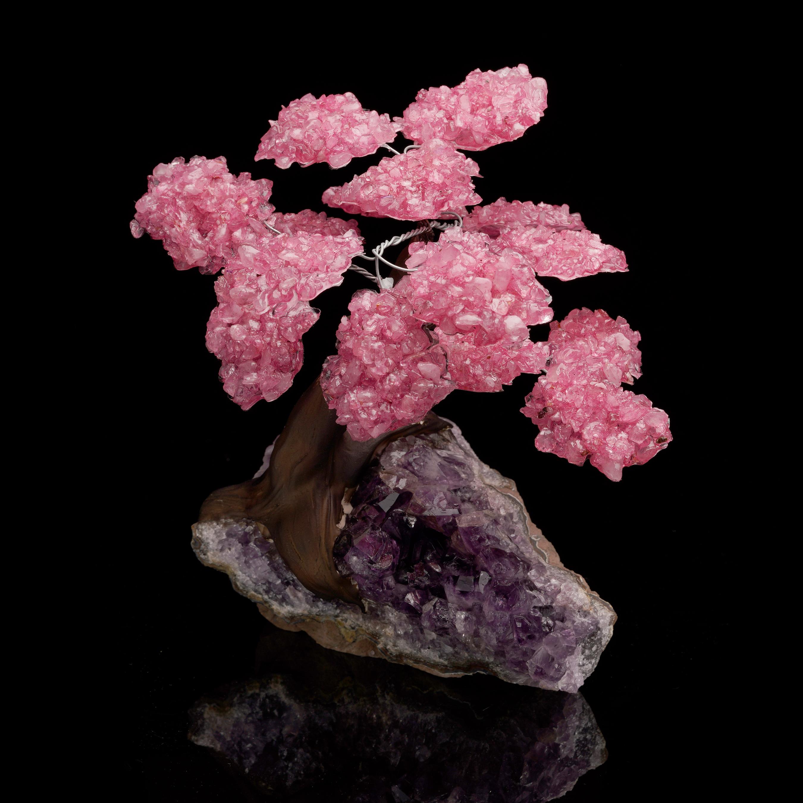 Dieser handgefertigte Edelsteinbaum zeigt rosa Rosenquarzkristallblüten, die an einem anmutig gewundenen dunklen Stamm erblühen, der elegant in einem natürlichen Amethystcluster verwurzelt ist. Der auffällige Kontrast zwischen den leuchtend