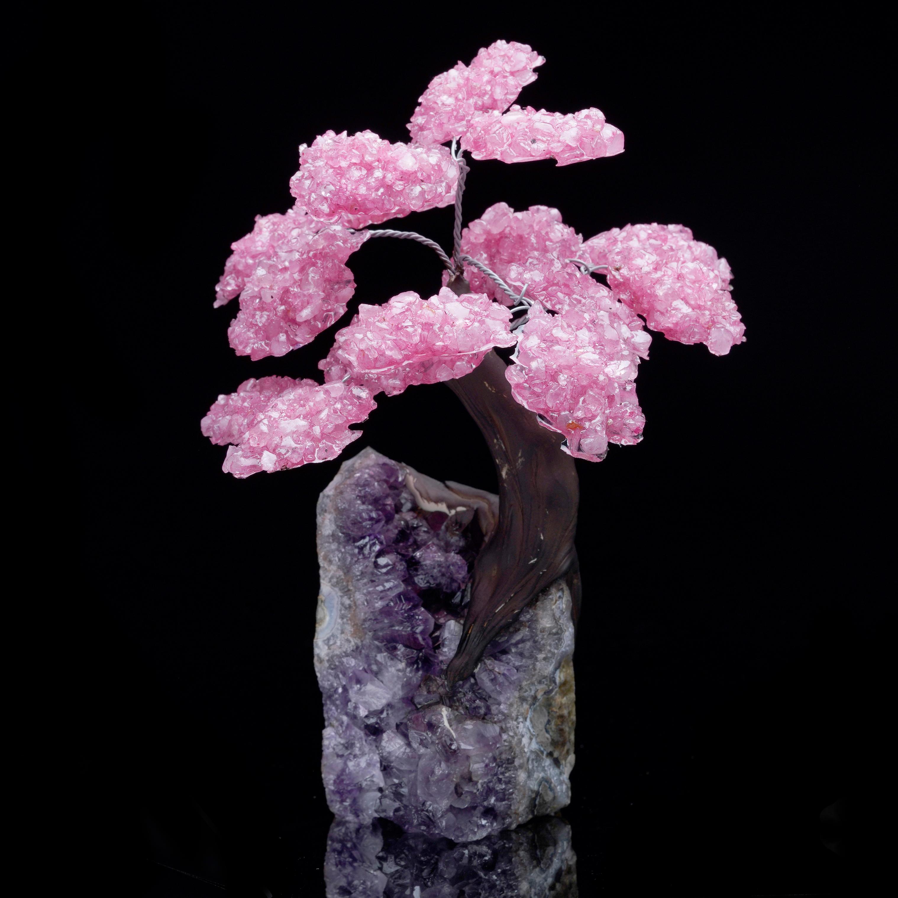 This handcrafted gemstone tree features pink Rose Quartz crystal blossoms blooming from a gracefully twisted dark trunk, all elegantly rooted in a natural Amethyst cluster base. The striking contrast between the vibrant pink Rose Quartz crystals and