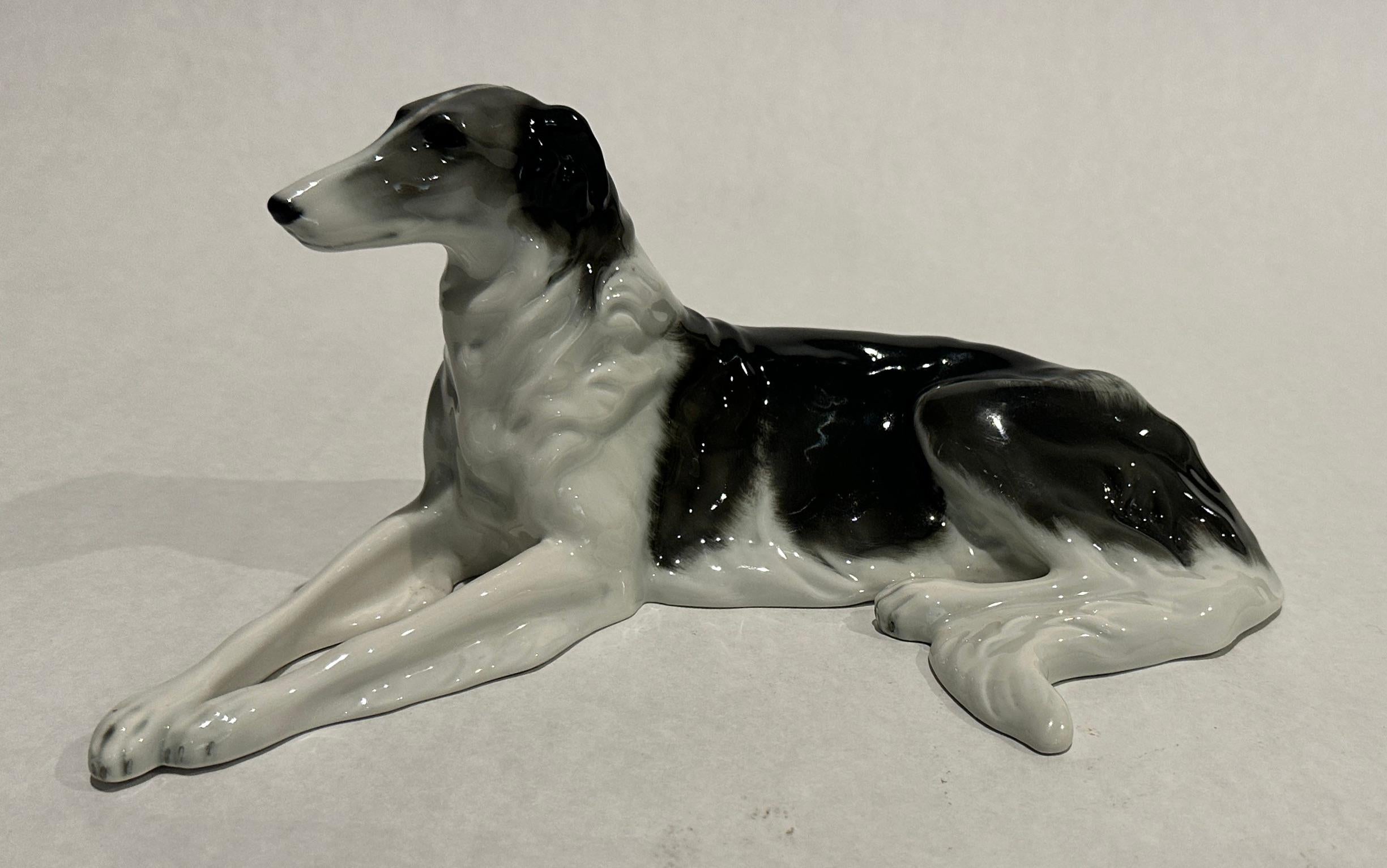Hundeskulptur aus Porzellan, Barsoi/Russischer Wolfshund, Darstellung eines auf dem Boden liegenden Barsois mit ausgestreckten Vorderbeinen, naturalistisch polychrom bemalt, auf der Rückseite über dem Standrand signiert. Diese detailreiche