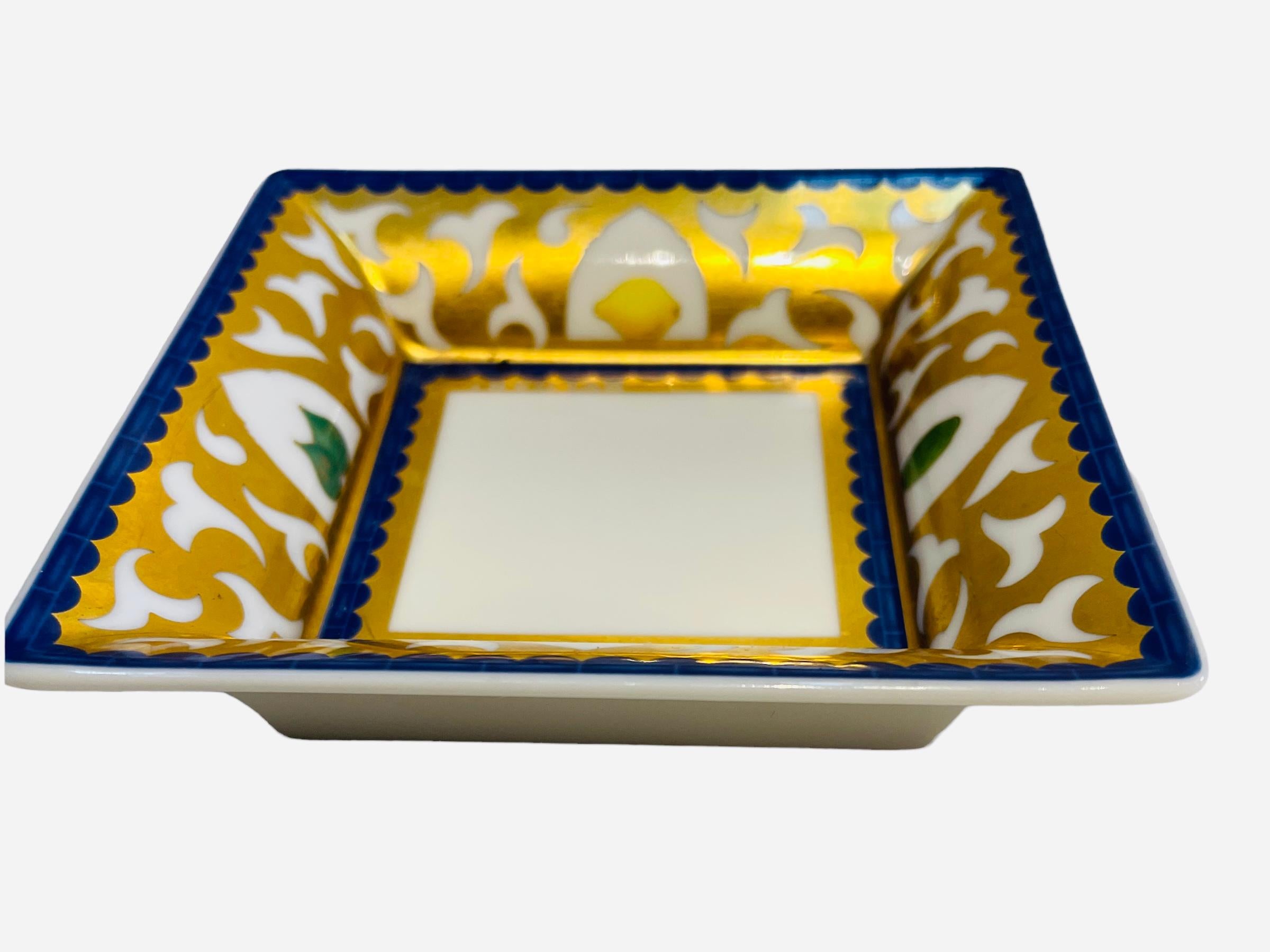 Se trata de un Plato decorativo de porcelana Rosenthal diseñado por Bulgari. Representa un plato cuadrado con fondo blanco y pintado a mano en color dorado y azul real alrededor de los lados. Cada lado del plato está decorado con un arco blanco que