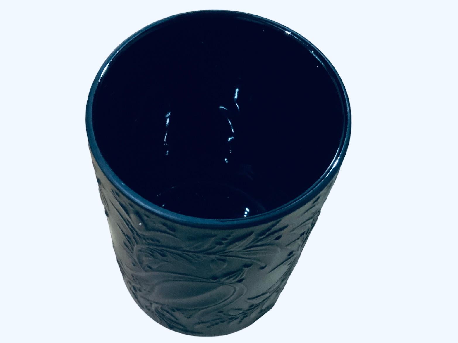 This is a Rosenthal Studio Bjorn Winblad Black Porcelain Small Vase. It depicts a small cylindrical vase embossed around with some flowers and leaves. Below the base is hallmarked Rosenthal Studio and signed by Bjorn Winblad.