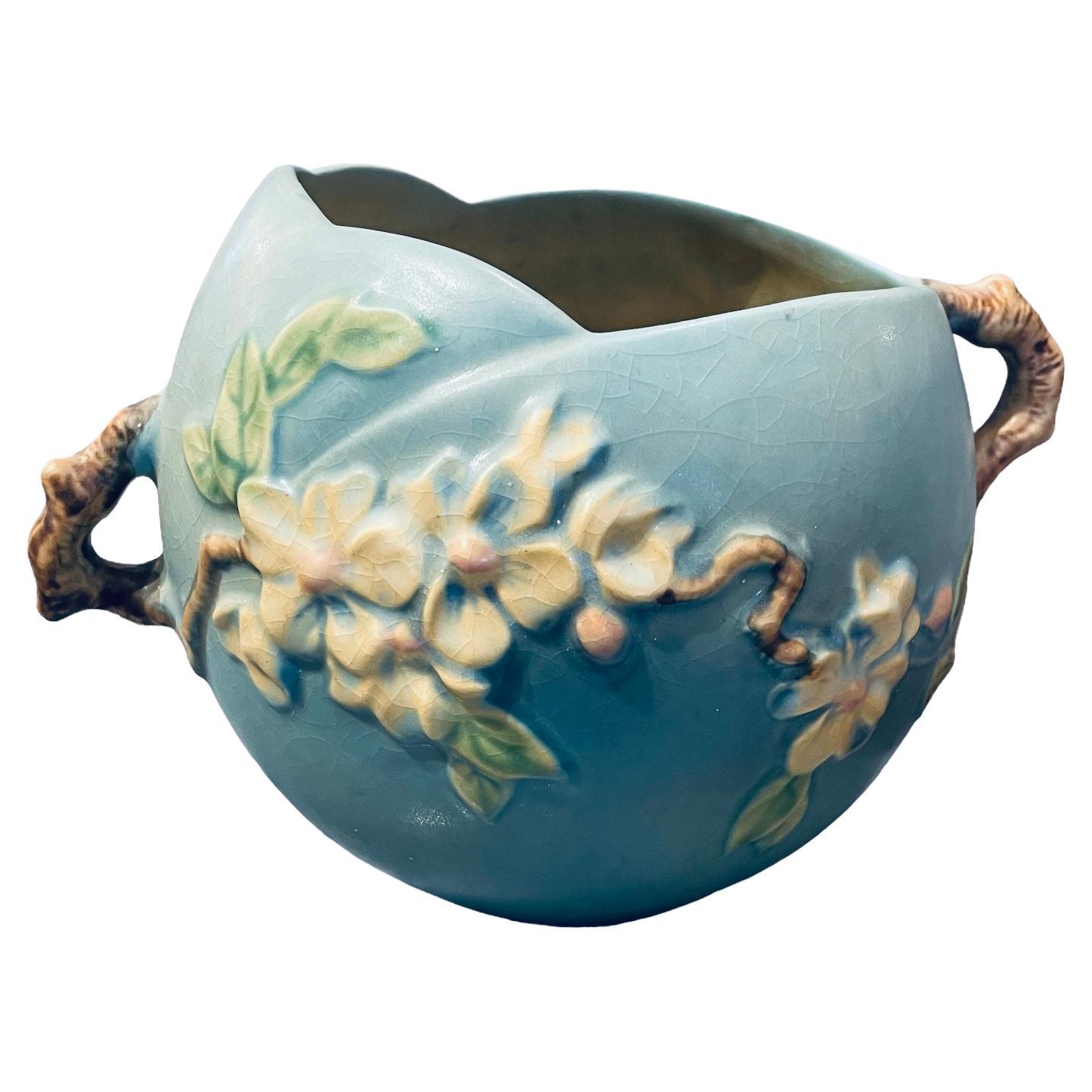 This is a Roseville Art Pottery Apple Blossom flowers pattern bowl. The bowl is blue and adorned with branches of white Apple Blossom flowers and green leaves in the front. Part of the branch made the handles at each side. Also, the upper border of