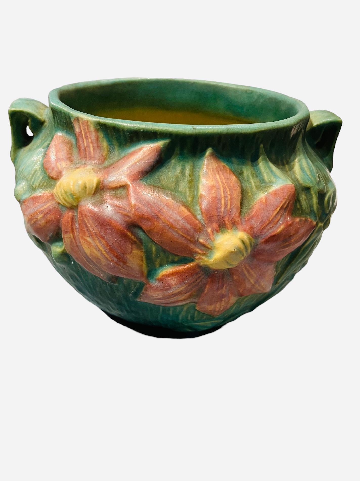 This is a Roseville Art Pottery Clematis flowers pattern bowl. The bowl is green and adorned with Clematis brown flowers with yellow center and green leaves in the front and back. Small triangular shaped handles are at each side of the body. Below