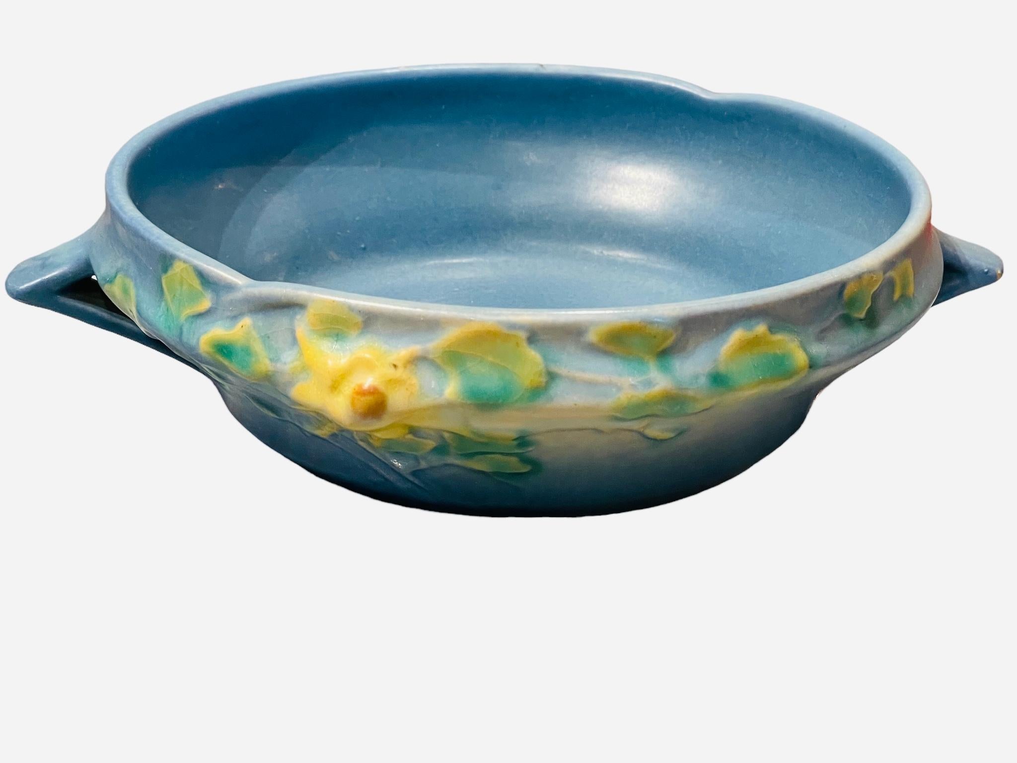 This is a Roseville Art Pottery Columbine pattern bowl. The bowl is blue and is adorned with a branch of yellow Columbine flowers and green/yellow leaves in the front. There are two triangular shaped handles at each side. Below the base is