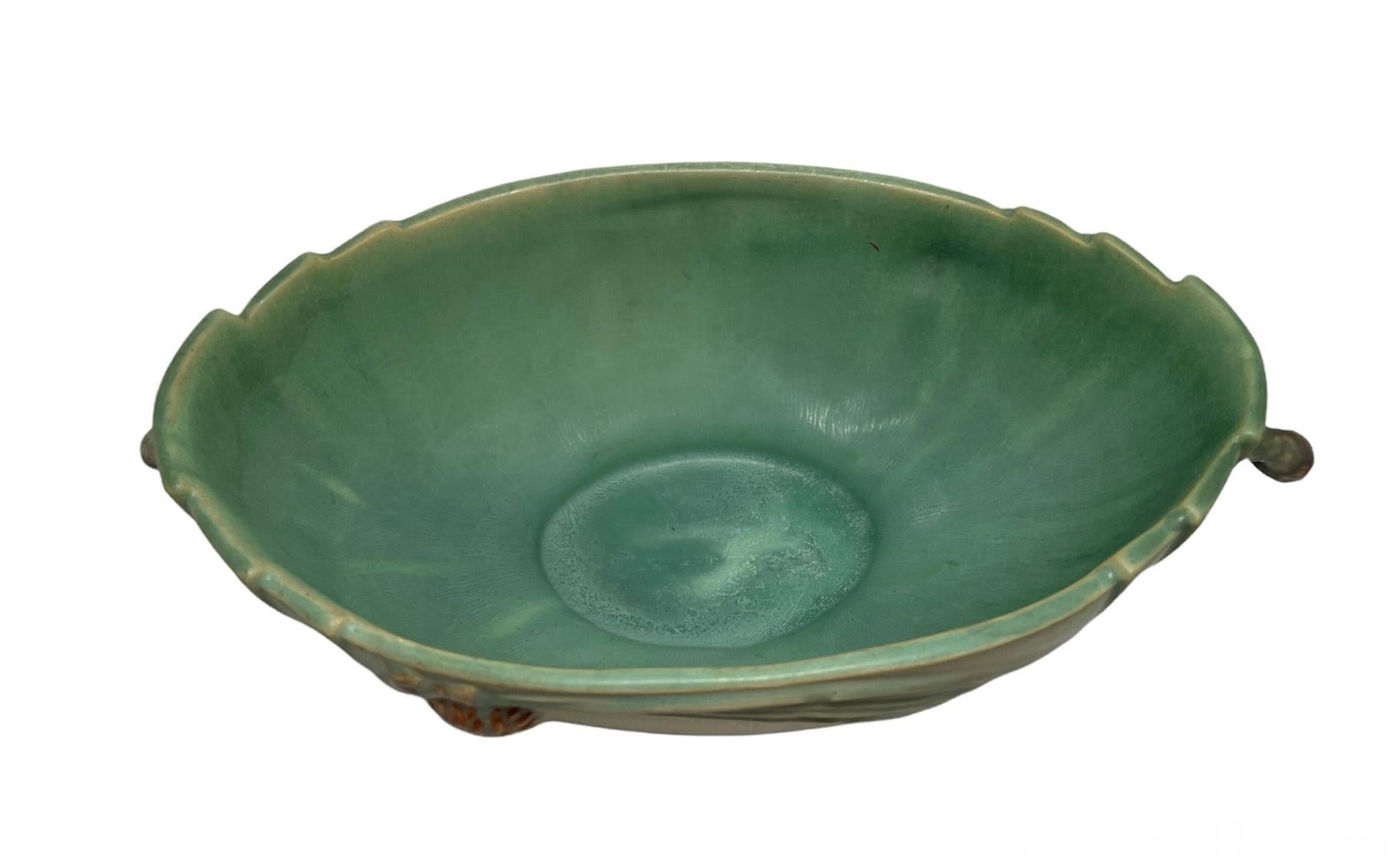 This is a Roseville Art Pottery Pine Cone pattern bowl. The bowl is green and is adorned with a branch of pine cone in the front. Part of the branch made the handles at each side. Also, each side of the bowl is reeded. Below the base is hallmarked