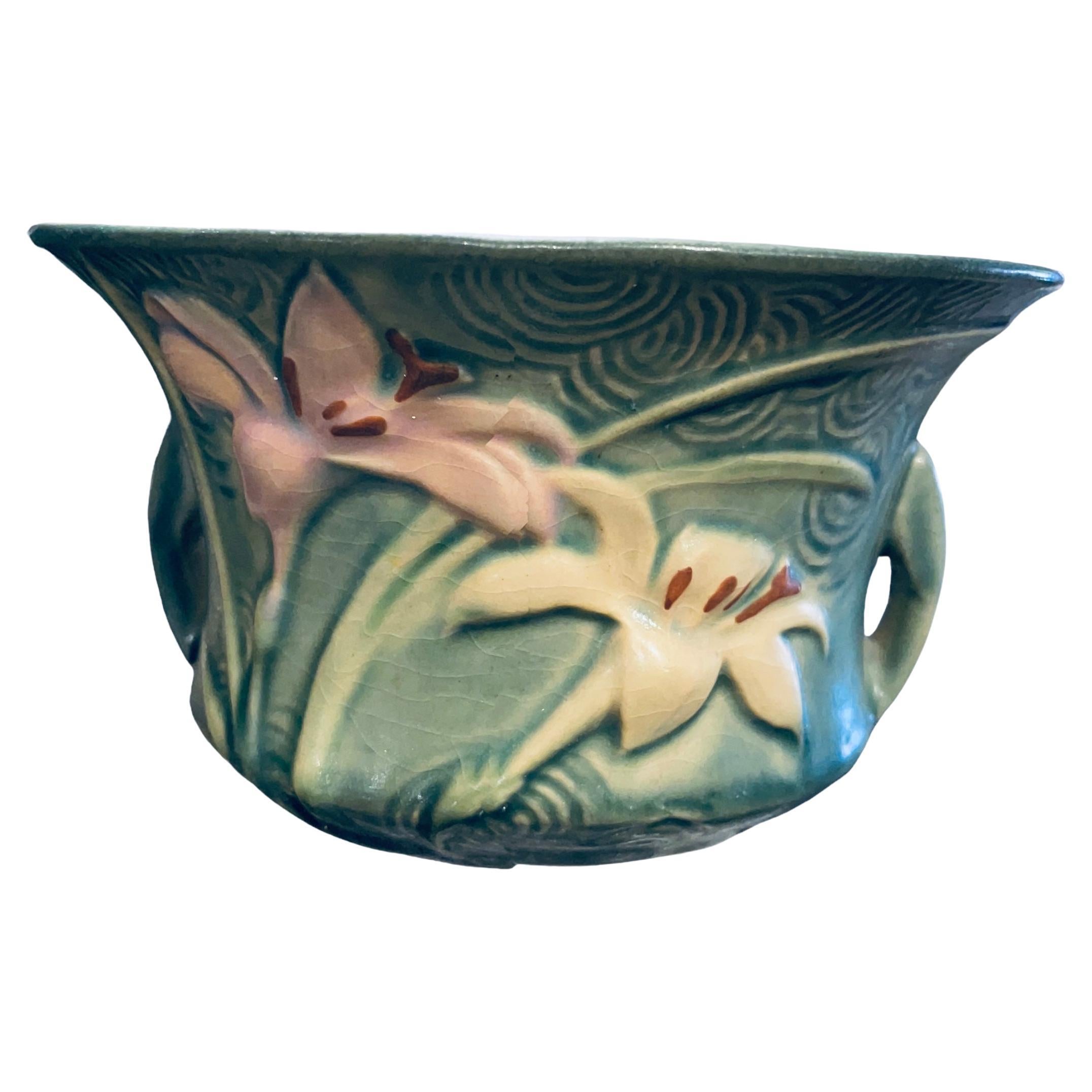 This is a Roseville Art Pottery Zephyr Lilies flowers pattern bowl. The bowl is bell shaped. It is green and adorned with Zephyr Lilies pink and white flowers with yellow/green leaves in the front and back. A relief of scalloped pattern also adorned