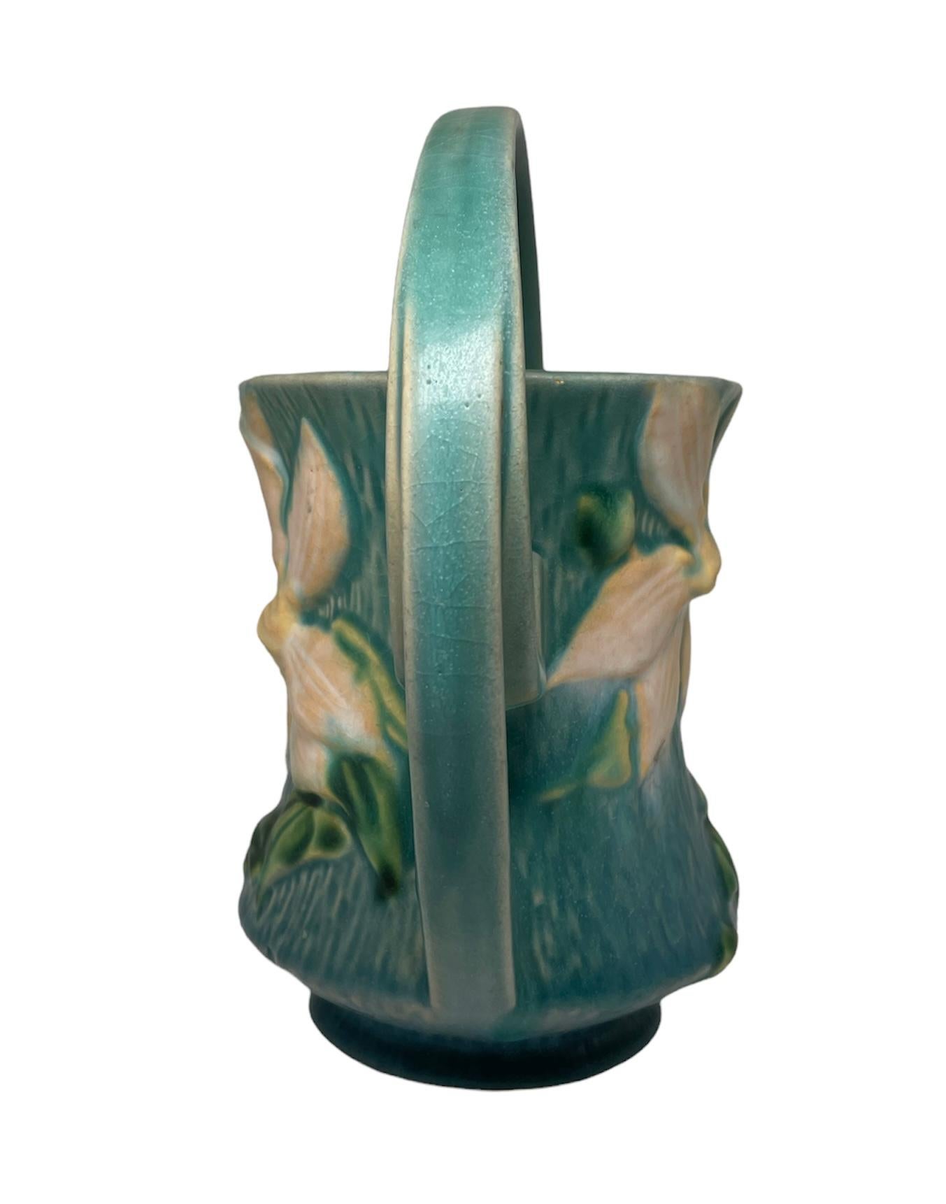 This a Roseville pottery art Clematis flower pattern basket. It depicts a green vase shaped as a basket and adorned with a large white Clematis flower in the center. At each side, there is a double handle that raise up forming a single arch. Below,