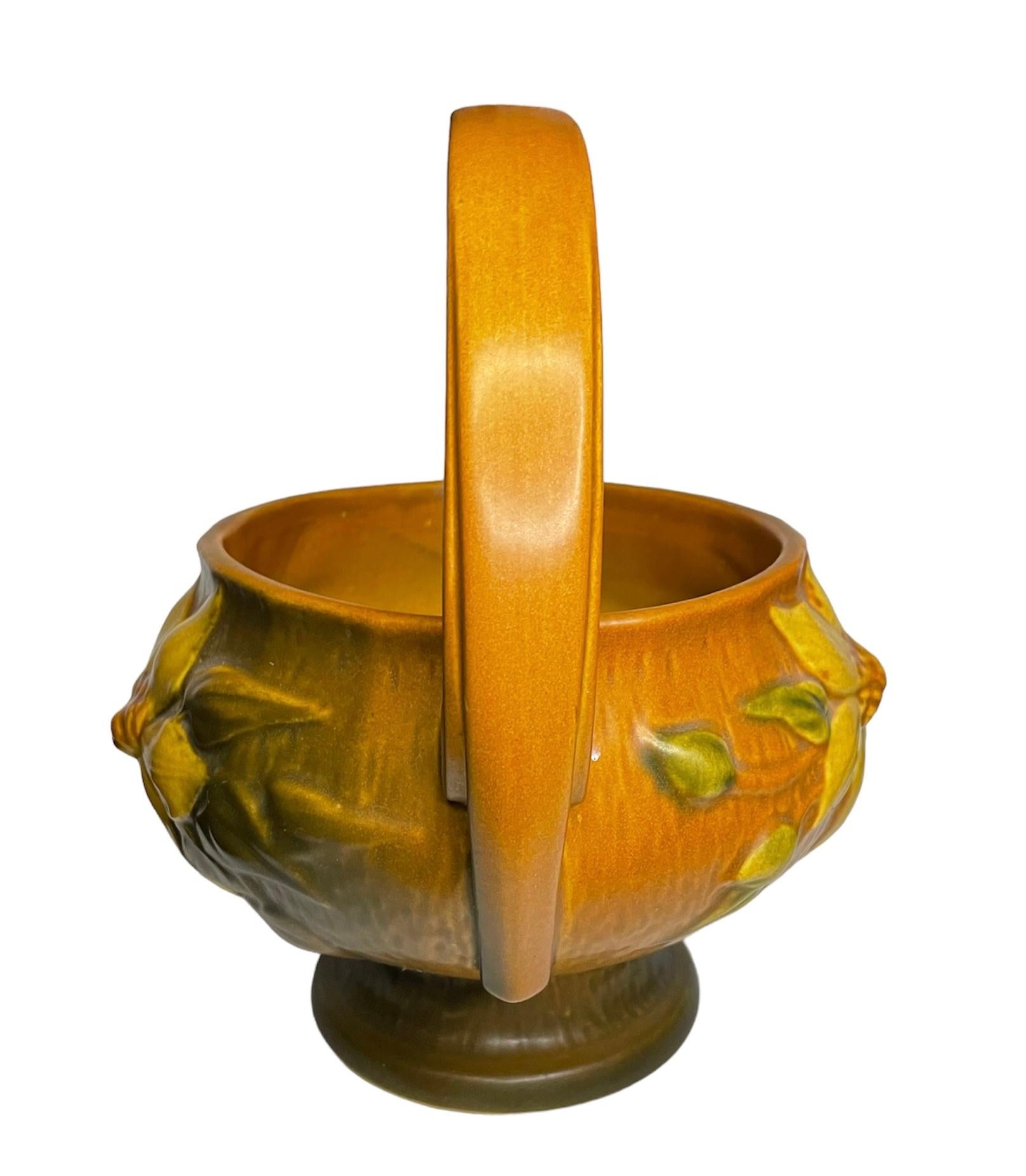 This a Roseville pottery art Clematis flower pattern basket. It depicts a brick color vase shaped as a round basket and adorned with two large yellow Clematis flowers in the front and one in the back center. At each side, there is a wide handle that