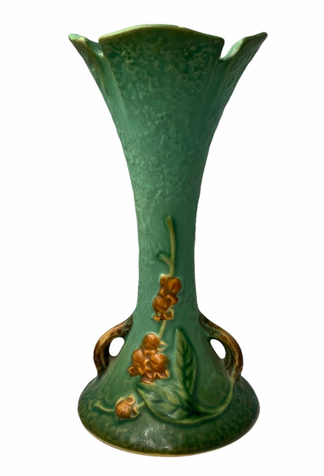 This is a green turquoise trumpet shaped Roseville vase. It is decorated with a relief of large green leaves & Bittersweet flowers. The upper border is shaped as flower’s petals. Two brown handles come out from the round base. Under the vase is