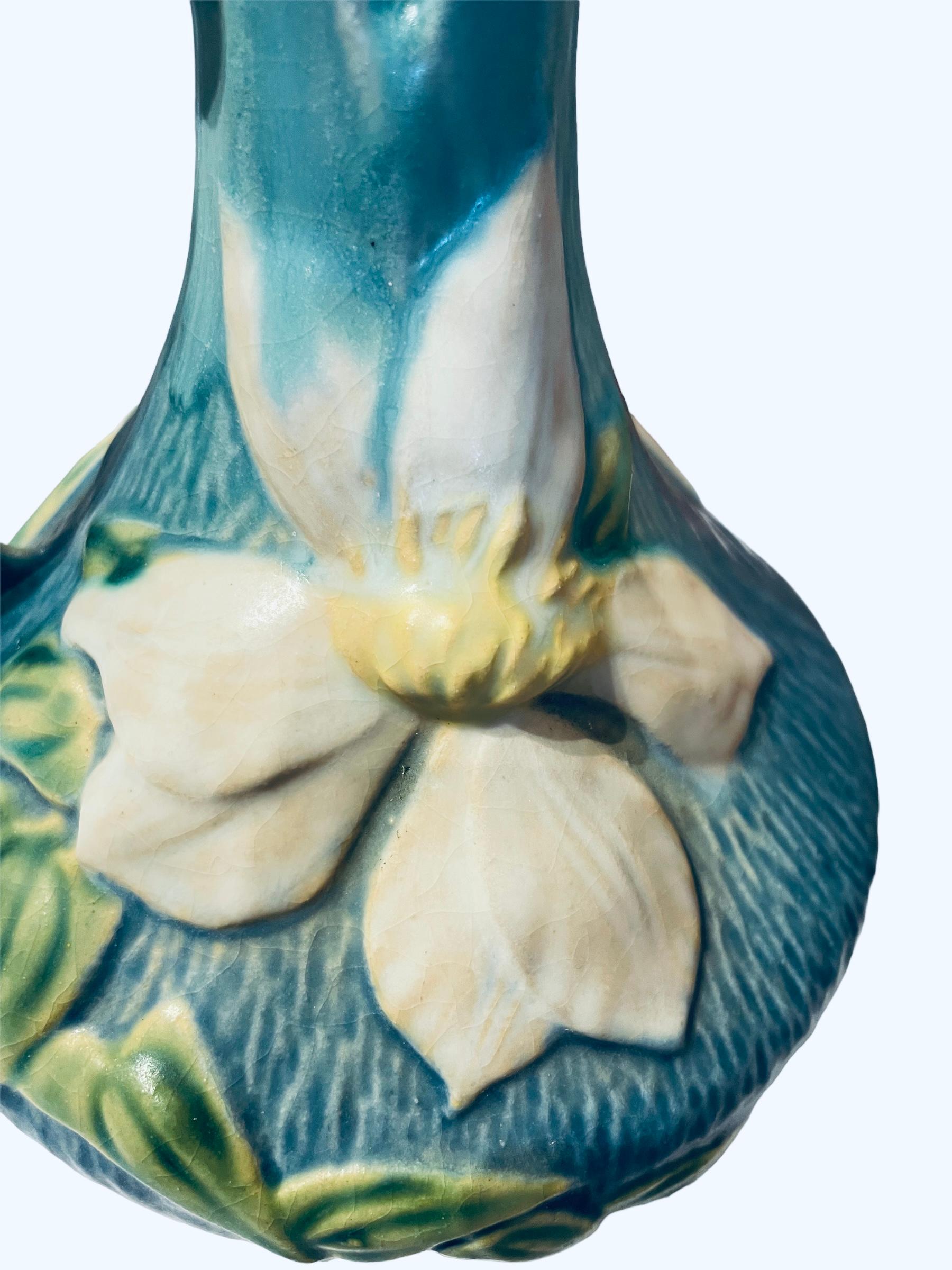 This is a blue turquoise jug shaped Roseville vase. It is decorated with a relief of large green leaves & white Clematis flowers. The upper border is shaped as flower’s petals. One handle come out from the neck to the body in one of the side. Under