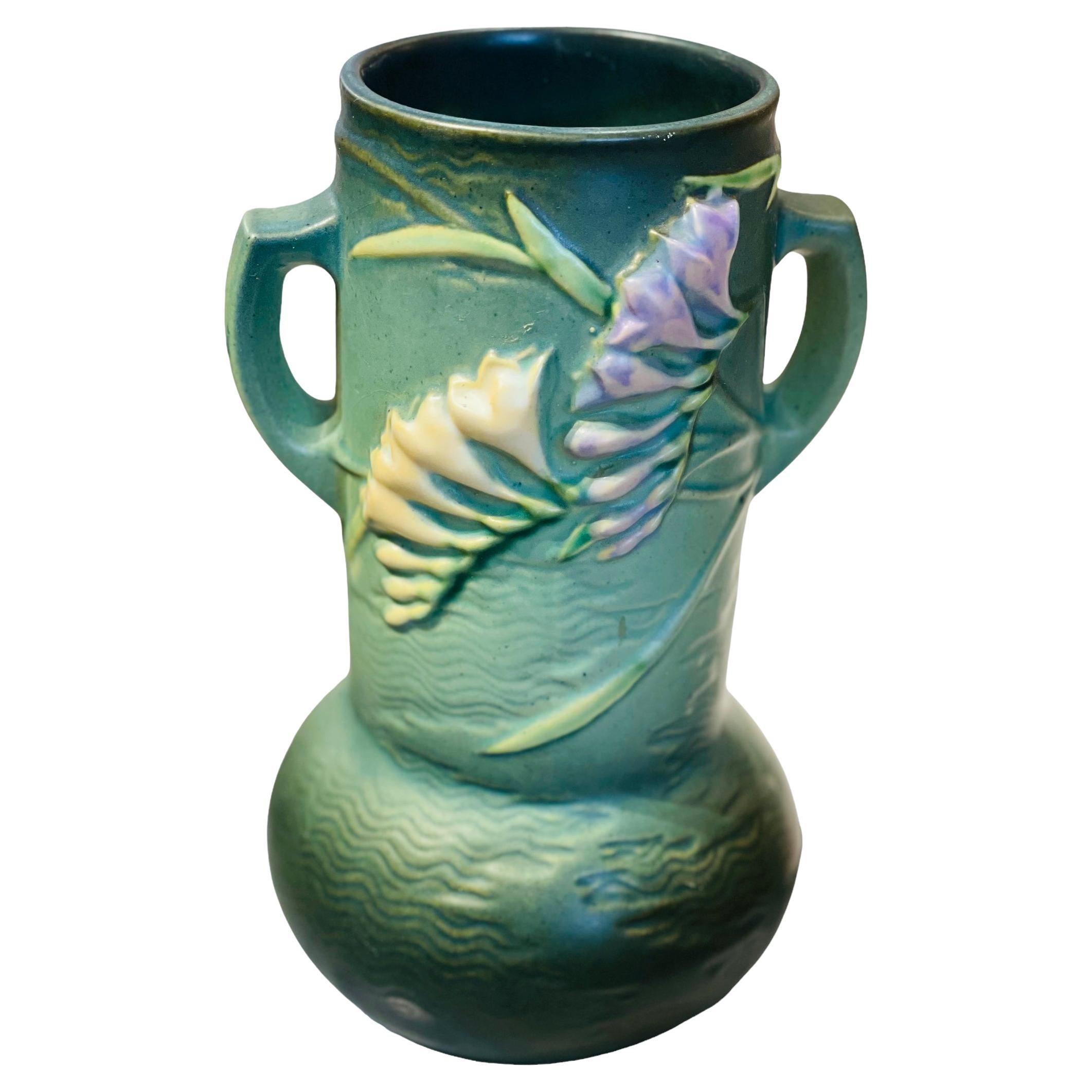 This is a green turquoise cylindrical bulbous shaped Roseville vase. It is decorated with a relief of long green leaves & branches of Freesia flowers in the back and front. The upper border is round shaped. Two D-shaped handles come out from the