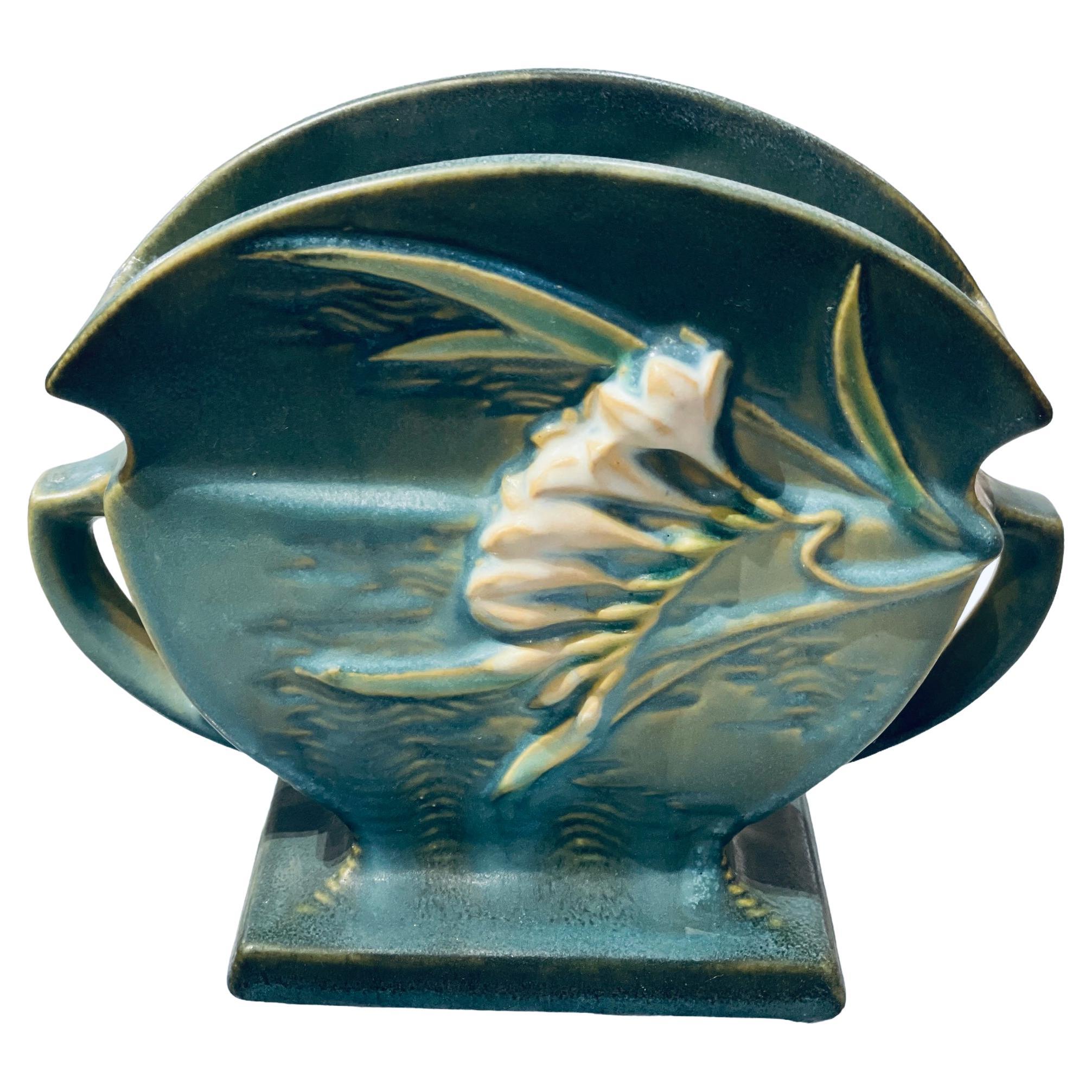 This is a green turquoise fan shaped Roseville vase. The fan shaped vase is attached to a rectangular base. It is decorated with a relief of long green yellow leaves & branches of Freesia purple and white flowers in the front and back. The upper