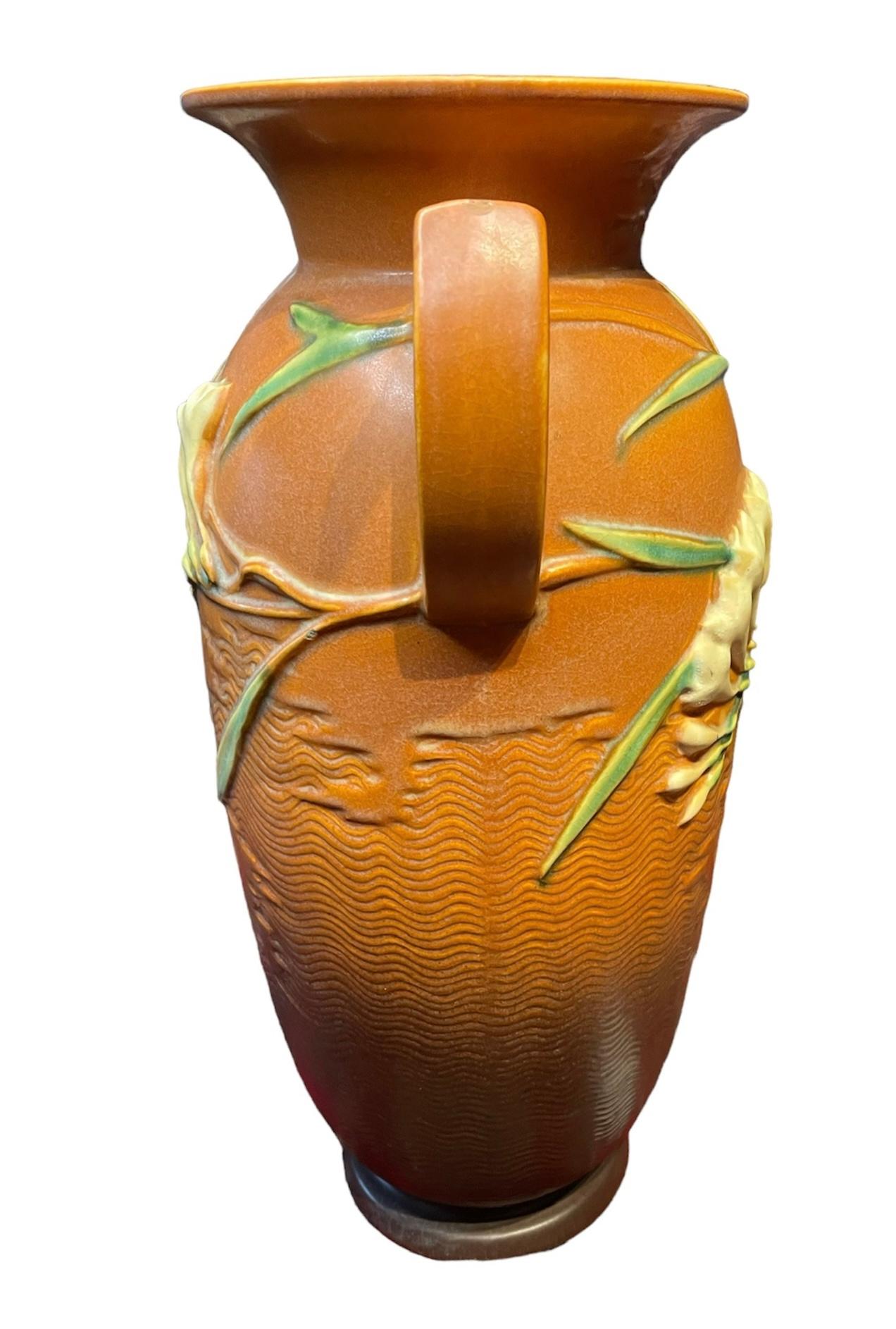 This is a brick/clay color jar shaped Roseville vase. It is decorated with a relief of long green leaves & branches of Freesia white and yellow flowers in the back and front. Two D-shaped handles come out from each side of the upper body. Under the
