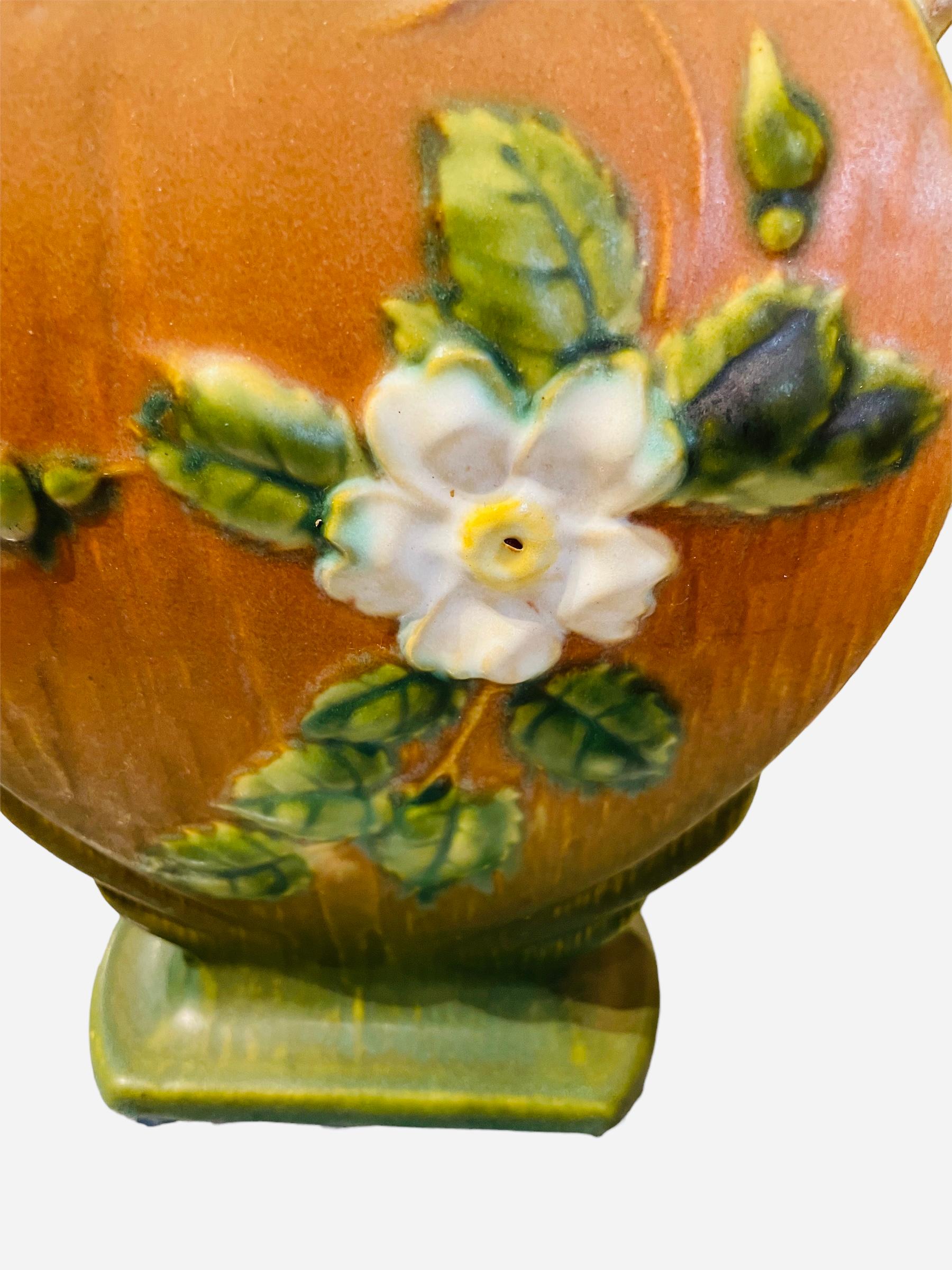 This is a Roseville Pottery White Rose flower heart shaped vase. The vase is green and brick color and adorned with a relief of White Roses flowers, green leaves and buds in the back and front. The upper rim is scalloped. The heart shaped vase is